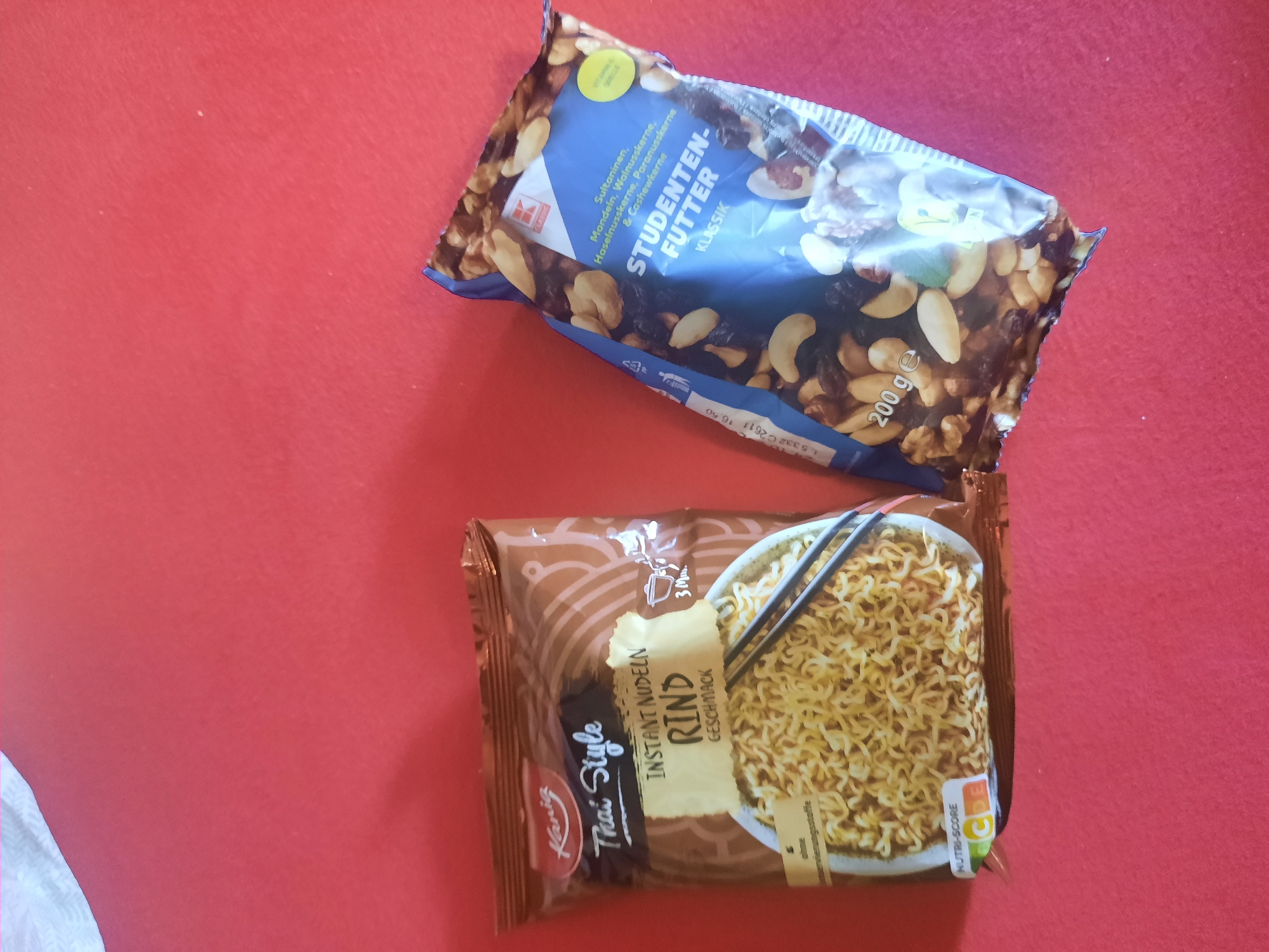 Two food product packages are displayed on a red, textured surface. On the left is a brown package of "Kania Thai Style Instant Nudeln Rind Geschmack" (Kania Thai Style Instant Noodles Beef Flavor). The package shows a bowl of noodles with chopsticks. On the right is a blue package of "STUDENTEN-FUTTER KLASSIK" (Student Food Classic), which appears to be a mix of nuts and dried fruit. The package lists ingredients such as sultanas, almonds, walnuts, hazelnuts, pecans, and cashews. The overall scene is a top-down, medium shot of these two items.