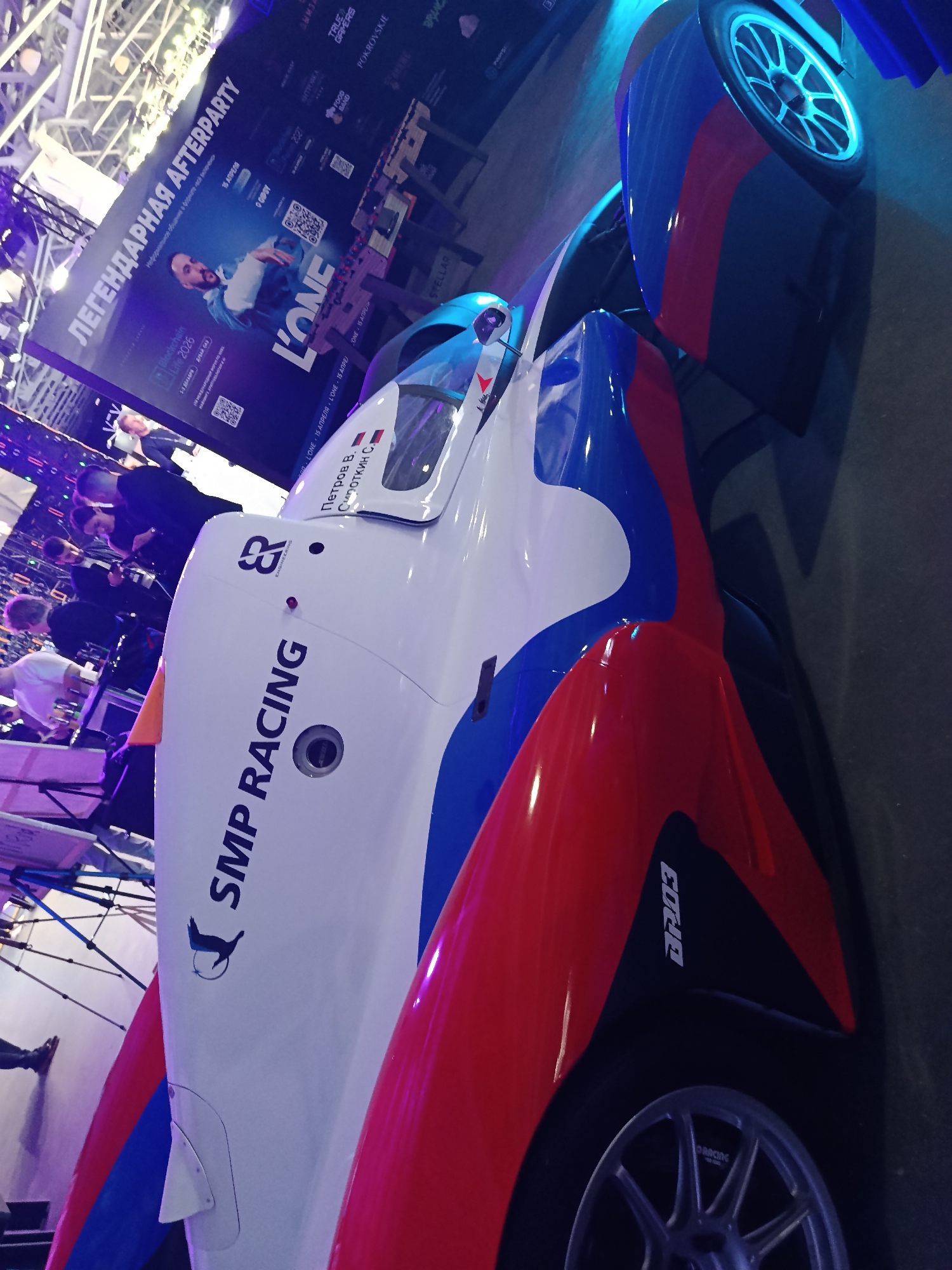 The image shows a racing car, emblazoned with "SMP RACING," with a red, white, and blue color scheme. The car is labeled with "Петров В. Сироткин С." A sign in the background advertises "ЛЕГЕНДАРНАЯ AFTERPARTY" and the "Blockchain Life 2026" event in Dubai, December 1-3. People can be seen blurred in the background, suggesting an event or exhibition setting. A catering table and stage are also visible.