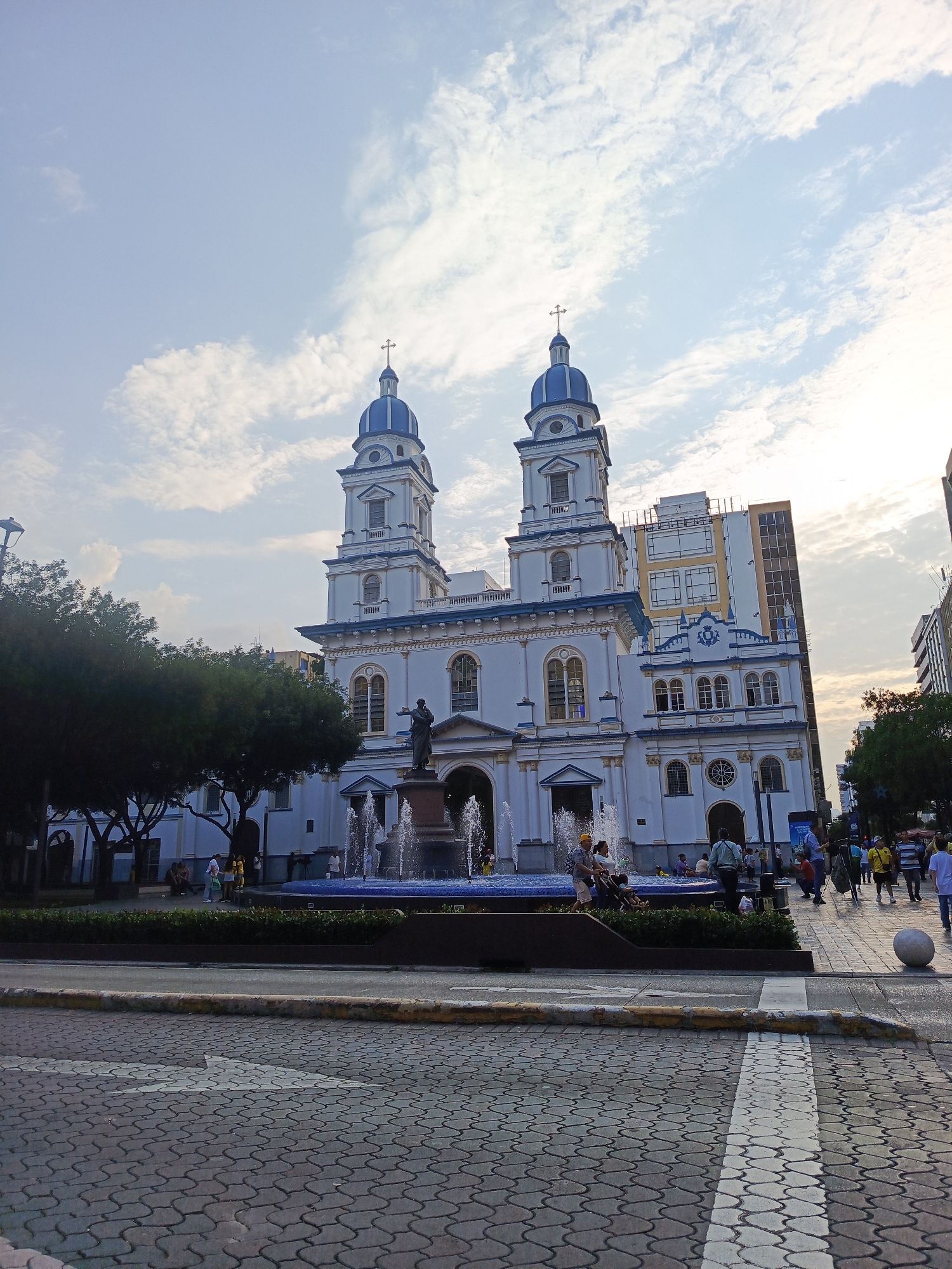 The image shows a white church with blue domes and accents, featuring two prominent towers topped with crosses. In front of the church, there is a fountain with water jets. A statue stands in front of the fountain. The church is situated in a public plaza with paved ground and some trees. Several people are visible in the plaza, walking and standing. In the background, there are modern buildings and more trees. The sky is partially cloudy. This scene is likely located in the historic district of Guayaquil, Ecuador, given the architectural style and the presence of a prominent church and public space.