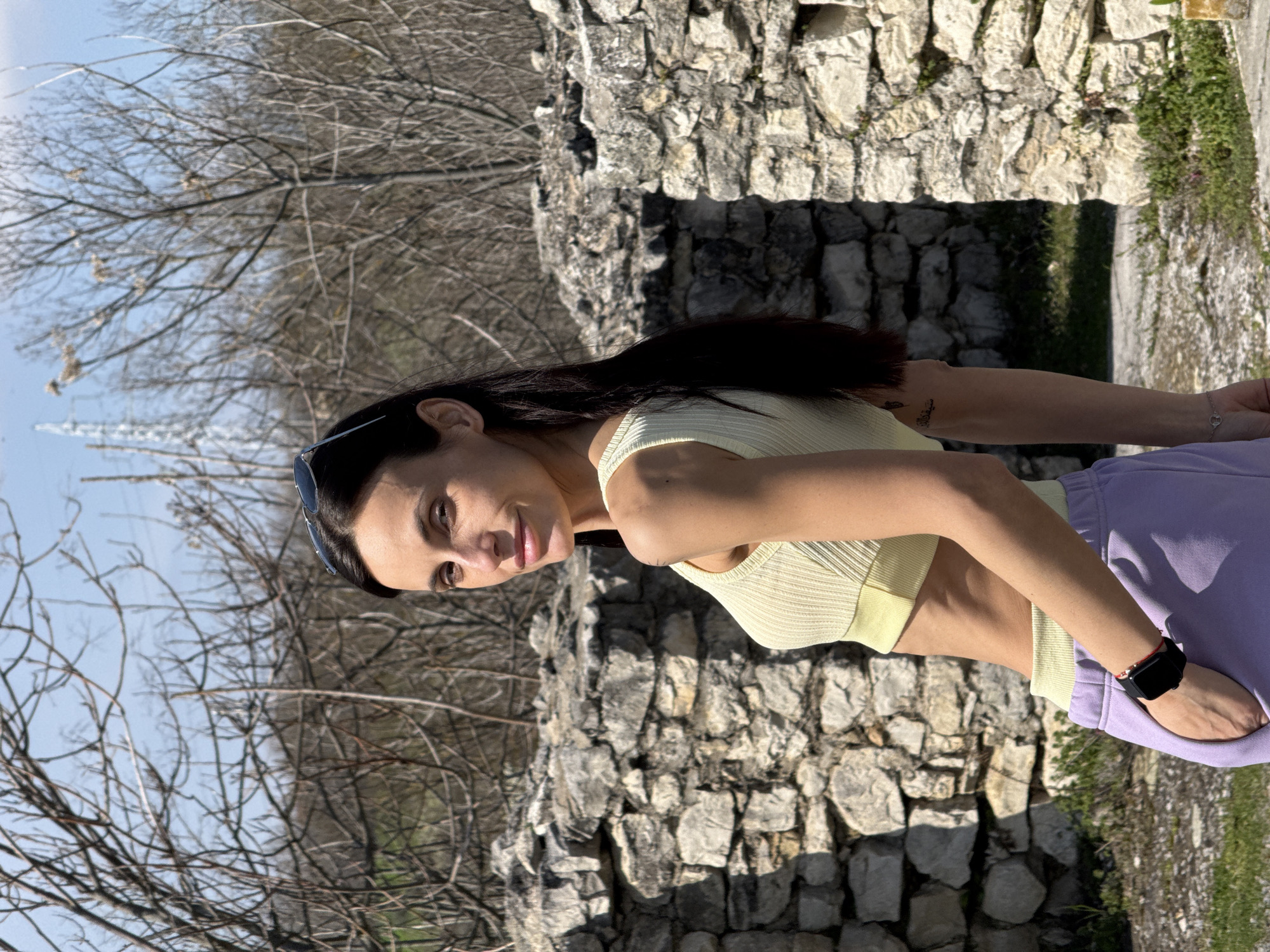 A woman stands outside in front of a stone structure. The woman has dark hair and is wearing a light-yellow tank top and light-purple sweatpants. She is wearing a watch on her right wrist. The stone structure is gray and appears to be an old wall or building. Behind the stone structure are trees with bare branches. The sky is visible in the upper left corner. The location may be in or near Pleven, Bulgaria, given the prompt.