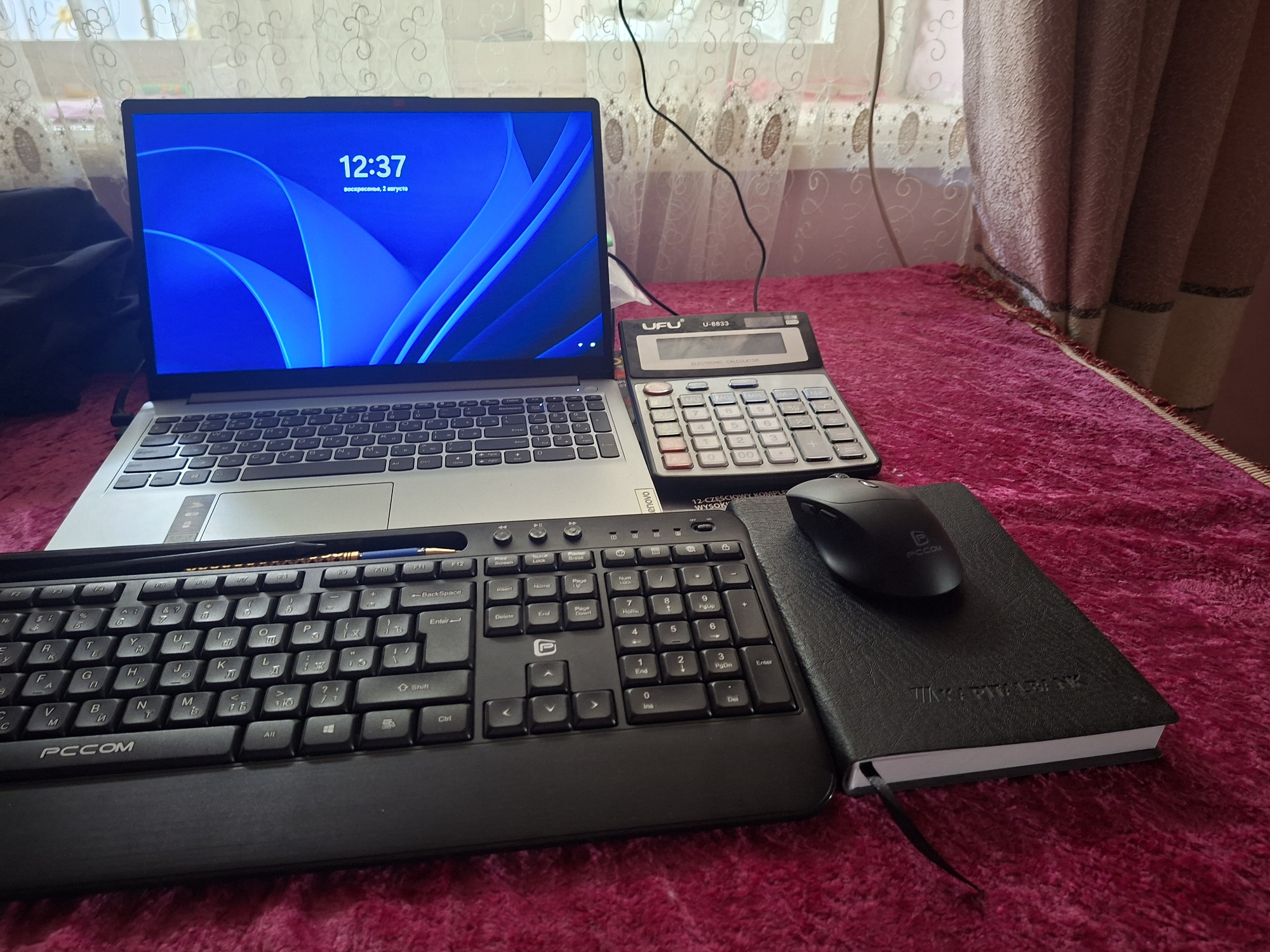 This is a medium shot, eye-level, indoor, still photograph of a workspace on a bed in Bustan, Uzbekistan. The objects in the scene are laid out on a plush, magenta-colored fabric.
Dominating the left side of the image is a silver and black laptop. The laptop screen displays a blue abstract wallpaper with the time "12:37" and text beneath it that reads "воскресенье, 2 августа." A black wired keyboard with Cyrillic lettering is positioned in front of the laptop, with the brand "PCCOM" visible on the lower left of the keyboard. A blue-tipped pen is resting on the top edge of the keyboard.
To the right of the laptop is a gray and black calculator, labeled "UFU U-8833" and "ELECTRONIC CALCULATOR." Next to the calculator, on the right side of the image, is a black notebook with the embossed text "ERITABANK" on the cover. A black computer mouse is placed on top of the notebook.
The background consists of a white sheer curtain with an intricate pattern, and a textured, light brown curtain. The lighting suggests it is daytime, with natural light coming from a window behind the curtains. There are no people visible in the image.