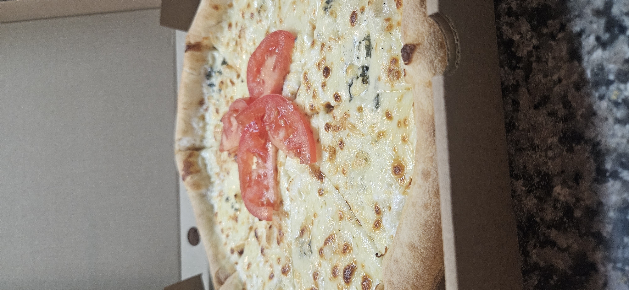 A partially-consumed pizza sits in a cardboard pizza box. Three slices of tomato sit atop the pizza. The pizza box rests on a speckled surface. The location of the image is not discernible from the visual details provided. It is not possible to identify the city of Sofia, Bulgaria or any associated details.