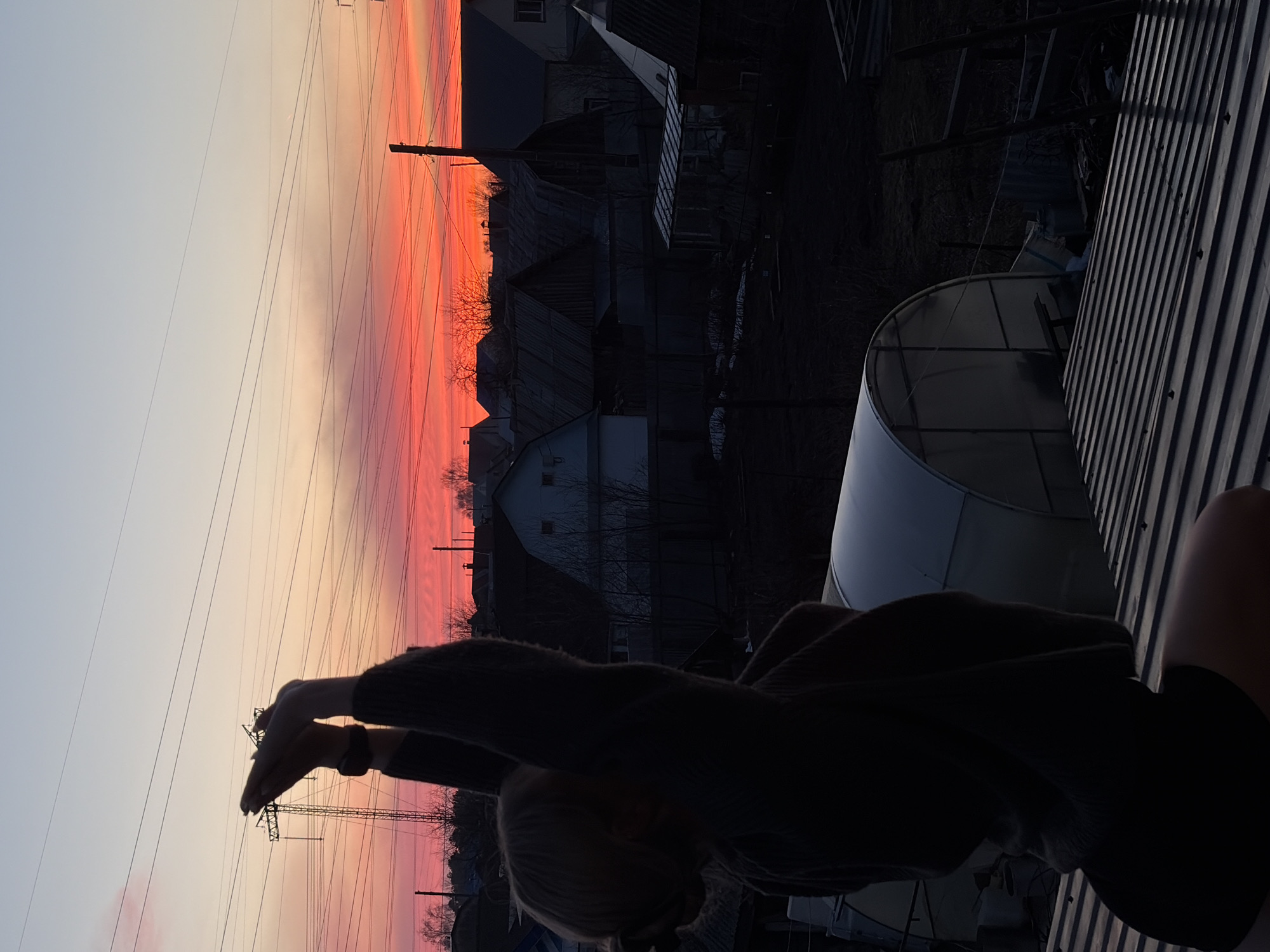 The image depicts a sunset scene from what appears to be a rooftop or elevated structure in Chaikovsky, Russia. The sky shows a gradient of colors, transitioning from a light peach/cream at the top to a deep orange/red near the horizon. Silhouetted against the sunset are buildings, possibly houses, and power lines with transmission towers. In the foreground, there is a dark, indistinct figure with their arms raised, seemingly interacting with the sunset. To the right, corrugated metal roofing and a semi-circular structure, possibly a greenhouse, are visible.