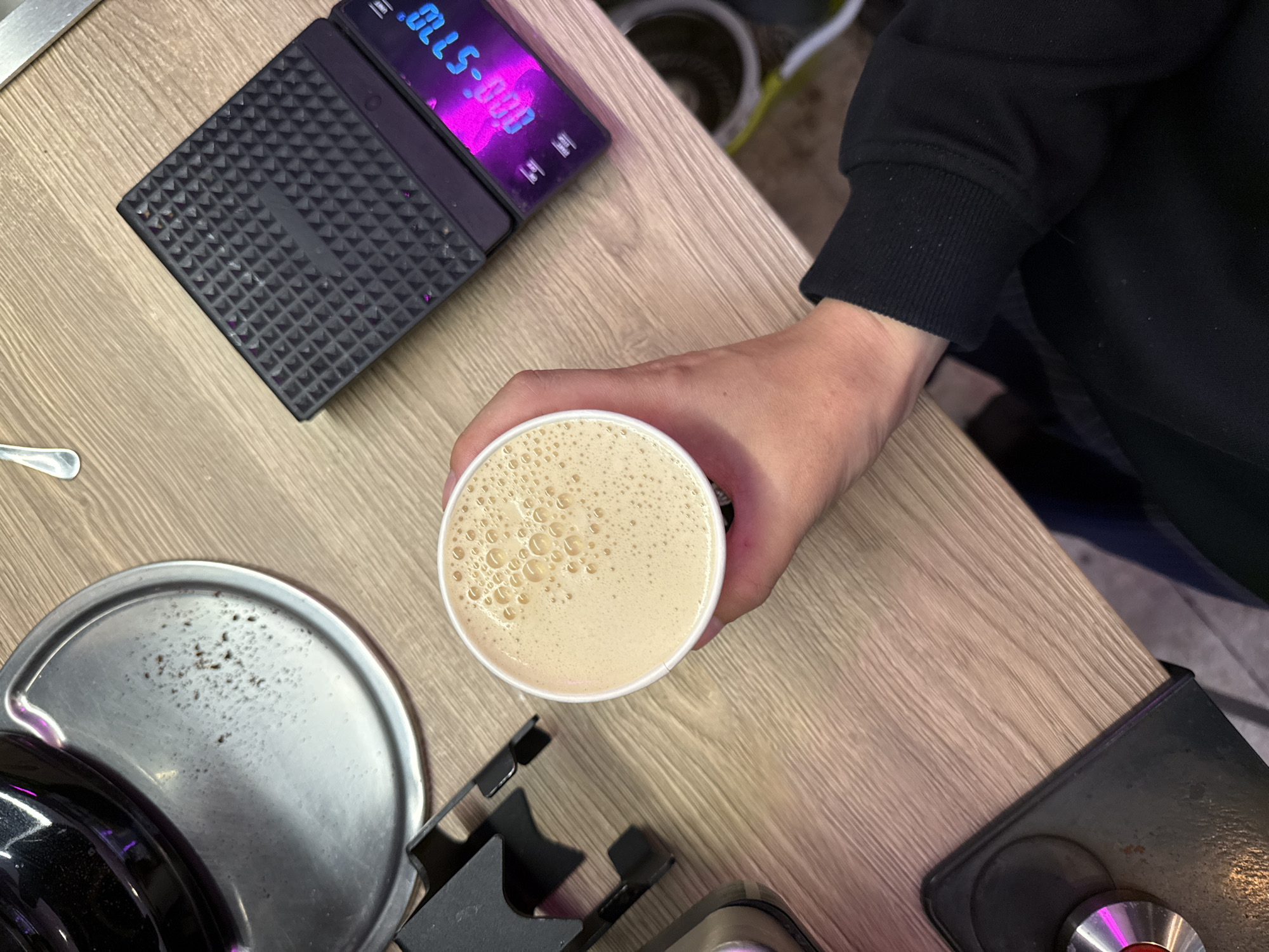 A top-down view of a person holding a paper cup filled with a foamy, beige-colored beverage. The cup is in the right hand of a person wearing a black sweatshirt. To the right of the cup is a black digital scale with a textured surface, displaying a magenta readout. To the left of the cup is a metal tray with a black handle and some dark specks on its surface. Further to the left is a black metal object with a holder. The scene is set on a light-colored wooden surface. The image does not provide any information that would allow for determining its location in Moscow, Russia.