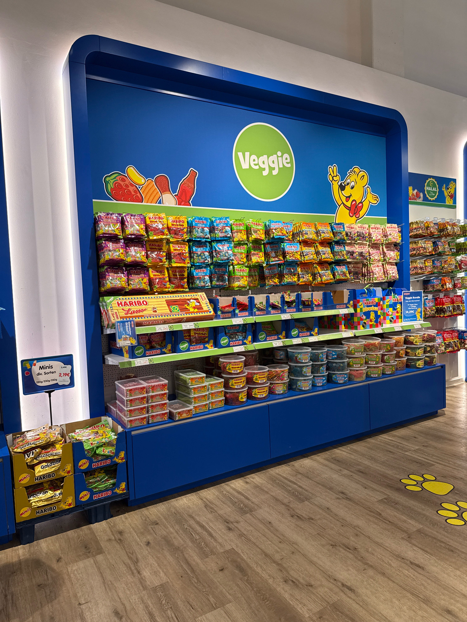 The image shows a brightly lit confectionery store display featuring a wide selection of Haribo candies. The display is predominantly blue and white, with vibrant signage and product packaging. A large blue shelf unit is filled with rows of bagged and boxed candies. The top of the unit features a large blue graphic with "Veggie" in a green circle and an illustration of a gummy bear giving a peace sign. Smaller signs indicate "Halal."
On the left side of the display, there is a price tag for "Minis div. Sorten 2,19€" and a smaller display box containing individually wrapped mini bags of Haribo candies, such as Goldbären and La(b)er.
The shelves are stocked with various Haribo products, including larger bags of gummies, boxed candies like "Haribo Lusso," and smaller tubs of assorted candies. Towards the bottom, there are multiple clear plastic tubs filled with different types of gummy candies.
The floor is made of light-colored wood-look laminate flooring. Yellow paw print decals are visible on the floor in the foreground, suggesting a playful or child-friendly theme. The overall impression is a well-organized and visually appealing candy retail environment. The image does not contain any specific information that allows for precise localization to a street or building in Wolfsburg, Germany.