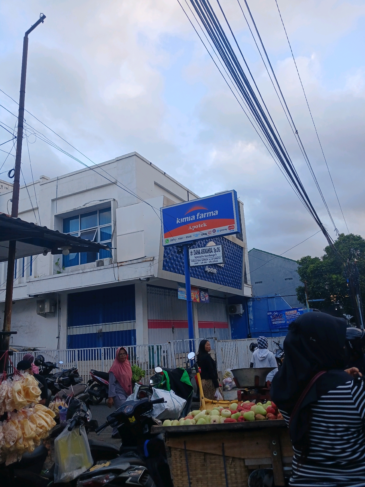 The image captures a street scene in Banyuwangi, Indonesia, under an overcast sky. In the foreground, several motorcycles are parked. To the left, a stand displays large bunches of light-colored snacks, possibly crackers or fried dough. In the center, a cart is laden with a variety of fruits, including apples and other red and green produce. A woman in a light pink hijab stands next to the motorcycles, and a woman in a black hijab with a striped shirt is in the lower right corner, partially obscuring the fruit cart.
Behind the motorcycles and fruit cart, a white building with blue shutters and windows dominates the scene. A prominent blue sign for "Kimia Farma Apotek" (a pharmacy chain) is mounted on the building. The sign also includes details about a doctor's practice, "DR. DIANA ARINANDA, Sp. OG." Further behind this building, another blue building is visible, and behind that, lush green trees can be seen. The sky is filled with heavy, grey clouds. Overhead, a dense network of dark electrical wires crisscrosses the frame, supported by a tall, dark pole on the left. A street lamp is attached to this pole. The overall impression is a busy urban environment with a blend of commercial activity and everyday life.