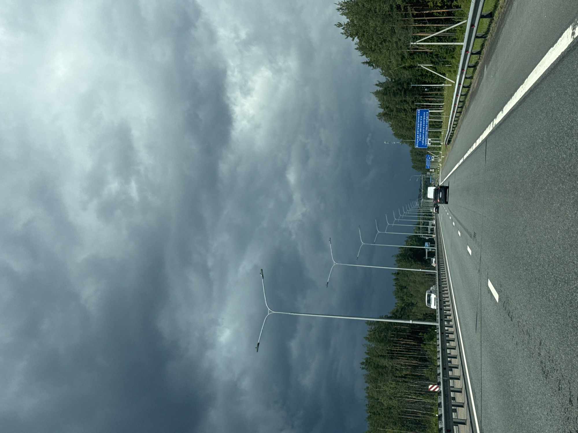The image shows a view from a vehicle traveling on a highway in Brilliant, Russia. The highway is paved with asphalt and marked with white lines. A metal guardrail separates the road from the adjacent forest. Tall trees, characteristic of a coniferous forest, line the side of the road.
Several streetlights are visible along the highway. In the distance, other vehicles, including cars and a white van, are also traveling on the road. A blue sign with Russian text is mounted on a post. The sign reads "Впереди автоматический контроль габаритного контроля транспортных средств," which translates to "Ahead automatic control of vehicle dimensions."
The sky is filled with dark, heavy clouds, suggesting an overcast or stormy day. The lighting is dim, indicative of a late afternoon or early evening time, possibly before a storm. The overall mood conveyed by the image is one of a journey through a potentially dramatic weather scenario, with the vastness of the sky and the looming clouds creating a sense of anticipation. The scene is a typical highway landscape, with the natural elements of the forest and the man-made elements of the road and its infrastructure.