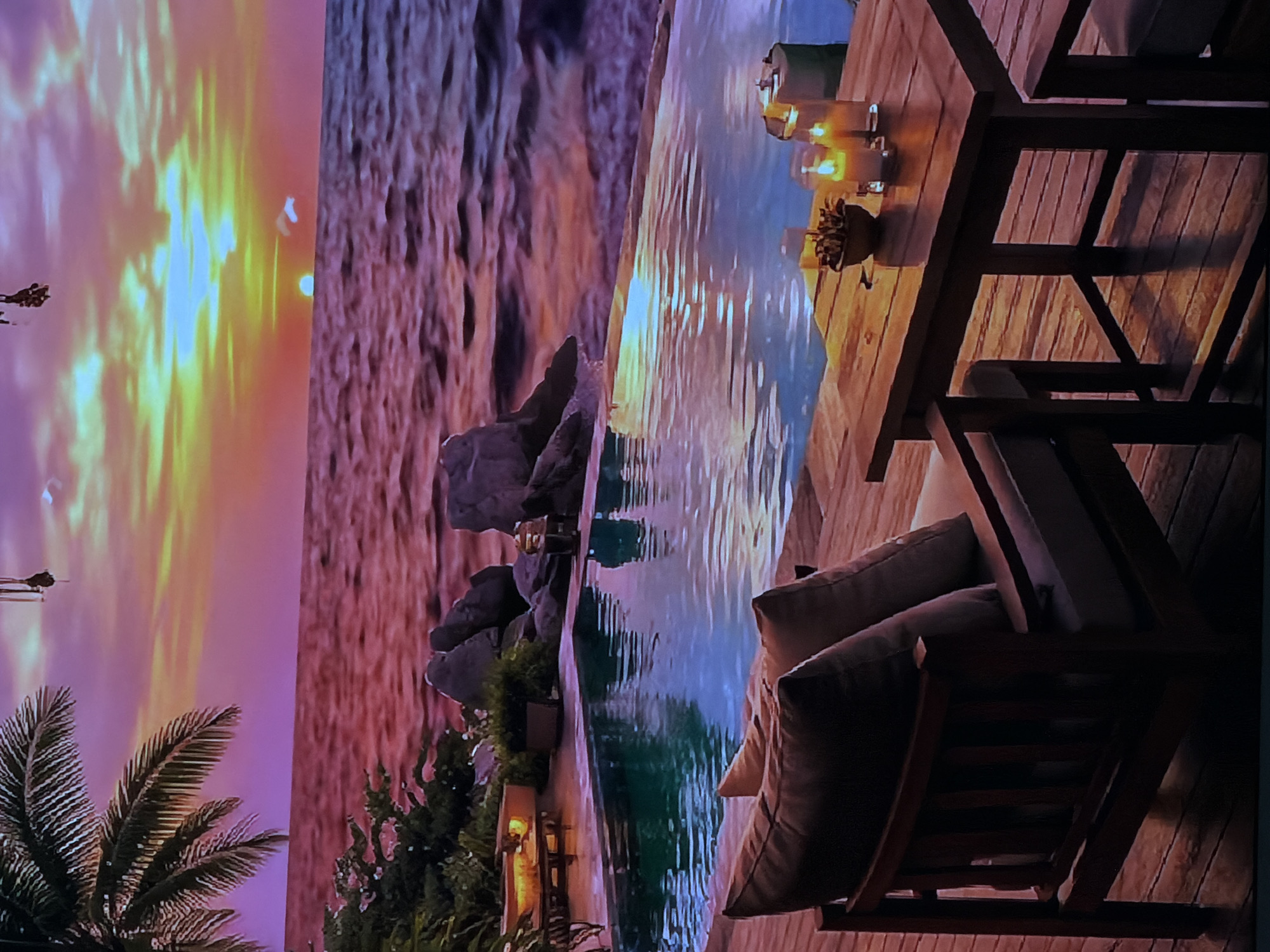 The image captures a serene and luxurious outdoor scene, likely at either sunset or sunrise. The sky on the left side of the frame displays a dramatic interplay of warm colors, transitioning from bright yellow and orange at the horizon to softer pinks and purples higher up, with reflections of these hues visible in the water. A large palm tree with visible fronds is partially seen in the lower left corner.
Adjacent to the sky is a textured, rugged landscape composed of rock formations, appearing in shades of brown and purplish-grey. Separating this landscape from the foreground is a calm body of water, likely a swimming pool or a decorative water feature. The water surface perfectly mirrors the vibrant colors of the sky, creating streaks of light and shadow. Several dark, prominent rocks are situated at the edge of the water, accompanied by small green bushes or plants.
On the right side of the image, a meticulously crafted wooden deck, featuring distinct planks, provides the setting for elegant outdoor furniture. A sturdy wooden table is adorned with multiple glowing lanterns or candles, casting a warm, inviting light. A small decorative plant or floral arrangement sits among the lights on the table. Comfortable-looking chairs or lounge seats, upholstered with thick, light-colored cushions, are partially visible, suggesting a space for relaxation. The furniture itself is made of dark wood, contrasting with the lighter tones of the deck planks.
No people or discernible text are visible in the image. The overall impression is one of tranquility, luxury, and natural beauty, evoking a resort-like ambiance ideal for relaxation. While the provided location context is Sofiivska Borshchahivka, Ukraine, the scene itself portrays a tropical or exotic setting.