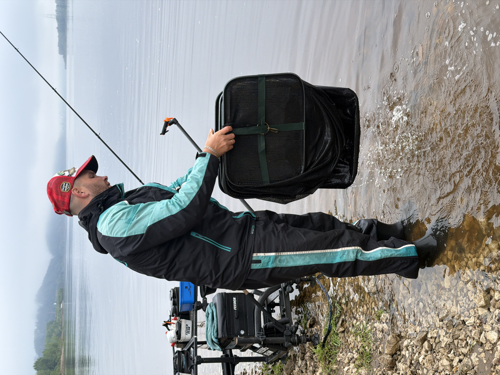 A man is engaged in fishing at the edge of a large body of water, possibly in Noviy, Russia. He stands at the water's edge, wearing black boots, black trousers with a teal stripe, and a black and teal/aqua two-toned jacket. On his head is a red and white baseball cap with "CATCH & RELEASE" written on it.
The man is actively handling a large, black fishing keepnet or landing net, which he is either deploying into or retrieving from the water. The net, secured by green straps, causes splashes and ripples in the calm, brown-toned water. A fishing rod extends diagonally into the frame to his left, indicating ongoing fishing activity.
Behind the man, on a stony and pebbly shore with some grass, is a comprehensive fishing setup. This includes a black box or seat prominently displaying "FEEDER" and "FEEDER TEAM" branding, along with a blue container and various other fishing accessories, all mounted on a stand or platform.
The scene is set outdoors on what appears to be an overcast day, providing soft, diffused light. The distant background shows faint outlines of land or mountains across the expansive body of water. The shoreline consists primarily of small rocks and pebbles.