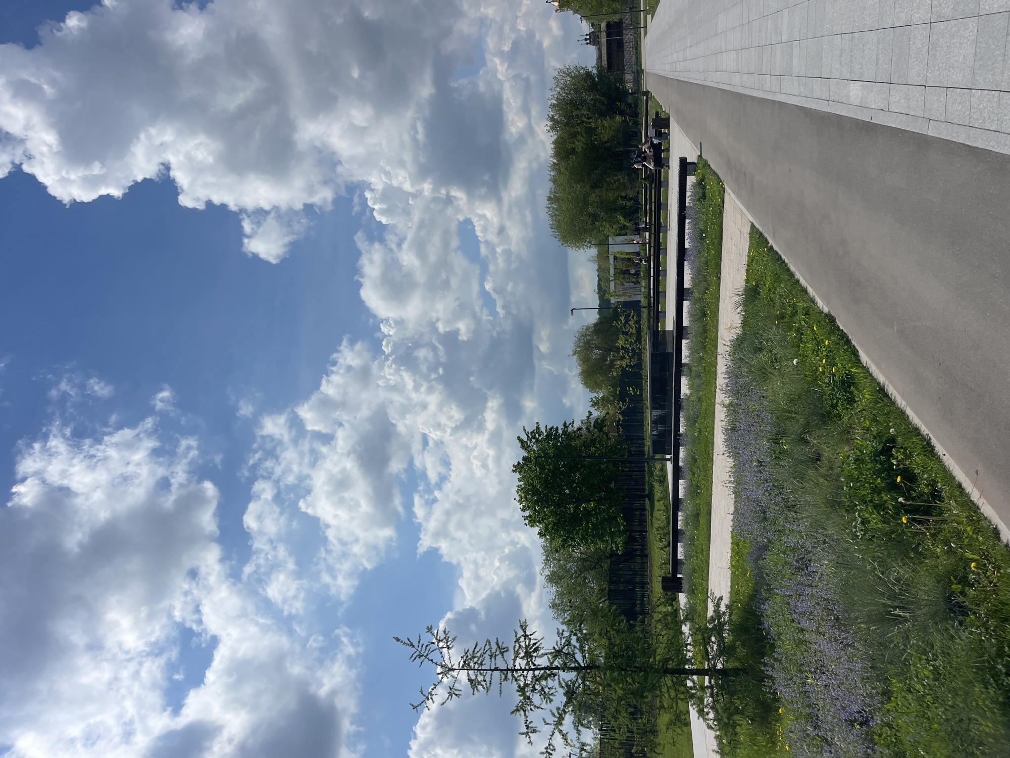 The image shows a park scene in Moscow, Russia. The sky occupies a large portion of the image and is filled with fluffy white clouds against a blue backdrop. On the right side of the image is a paved walkway or path, bordered by a strip of grass and flowering plants. A black metal fence runs parallel to the path, separating it from the greenery. Benches are placed along the fence. Trees are visible behind the fence, adding to the park-like setting. In the distance, some architectural structures can be faintly discerned. The scene appears to be taken during the daytime, under natural lighting conditions.