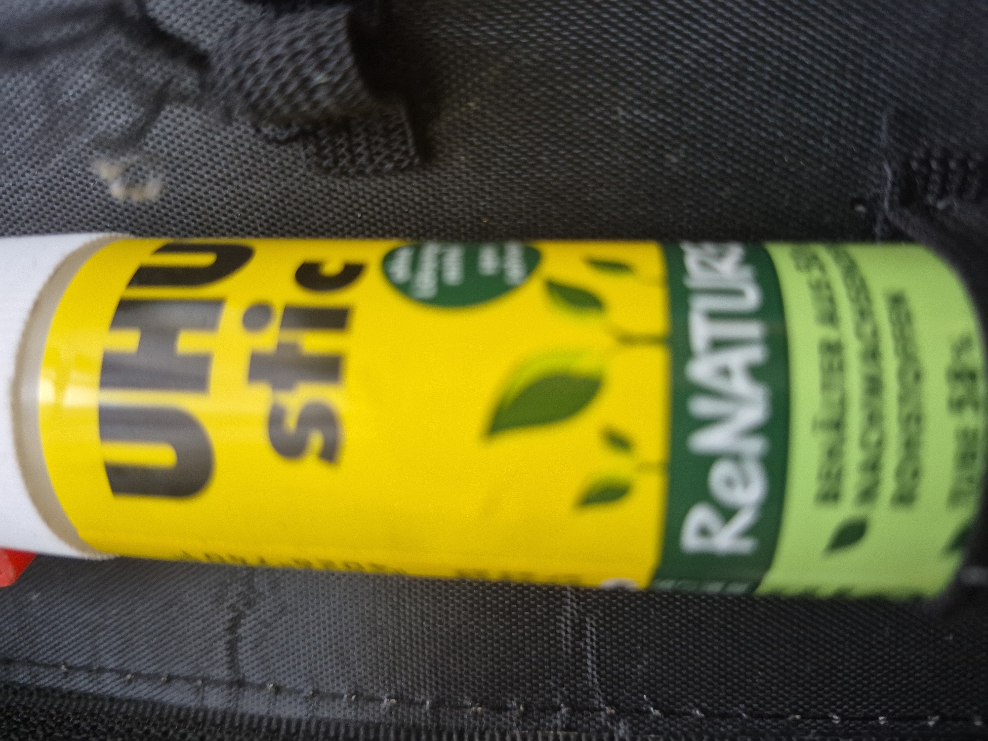 This media file is a close-up, eye-level shot of a UHU stic glue stick with a "ReNature" label, lying on a textured black surface, possibly fabric like nylon or polyester, which appears to be part of a bag or backpack due to the visible stitching. The glue stick is predominantly yellow with green accents.
The text visible on the glue stick includes: "UHU stic" in large black font on the yellow background. On the green portion, the text "ReNATURE" is prominently displayed in white. Below "ReNATURE", there is smaller text which appears to be in German. While blurry, some phrases can be partially deciphered, such as "BEHÄLTER AUS..." and "NACHHALTIGKEITS...", suggesting information about the container and sustainability. There is also a percentage visible, "58%". Small green leaf graphics are scattered on the green section, reinforcing the "ReNature" theme.
The lighting appears to be artificial and fairly uniform, with no strong shadows or indications of natural light. The background is out of focus, emphasizing the product in the foreground. There are no people or animals visible in the frame. The scene is static, with the object simply presented against the dark background. The overall impression is that of a product shot, highlighting the features of the UHU stic ReNature glue stick, particularly its eco-friendly aspect.