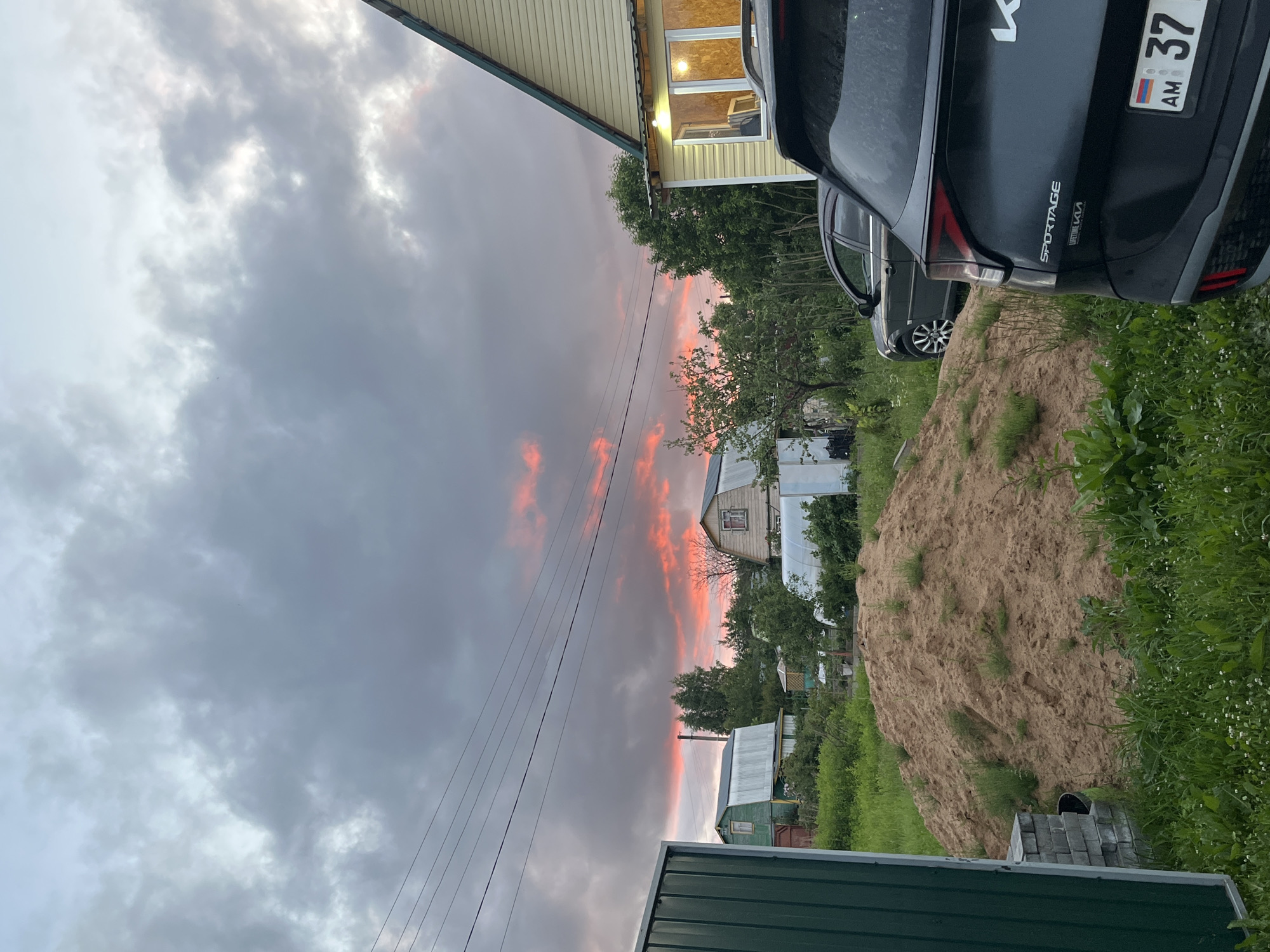The image captures a rural scene in the city of Klyobushnevo, Russia, at dusk. A vibrant sunset with bright pink and orange hues is visible in the stormy grey clouds. In the foreground, a large pile of sand and green grass dominate the ground. To the right, the rear of a dark gray Kia Sportage is parked, with its license plate displaying "AM 37". Behind the Kia, a dark sedan is visible. A yellow house with a green roof is situated on the right side of the frame, with lights on inside its windows. In the background, several small houses with metal roofs, trees, and a greenhouse are scattered across the landscape, along with power lines stretching across the sky. A green metal fence is partially visible on the left side of the image.
