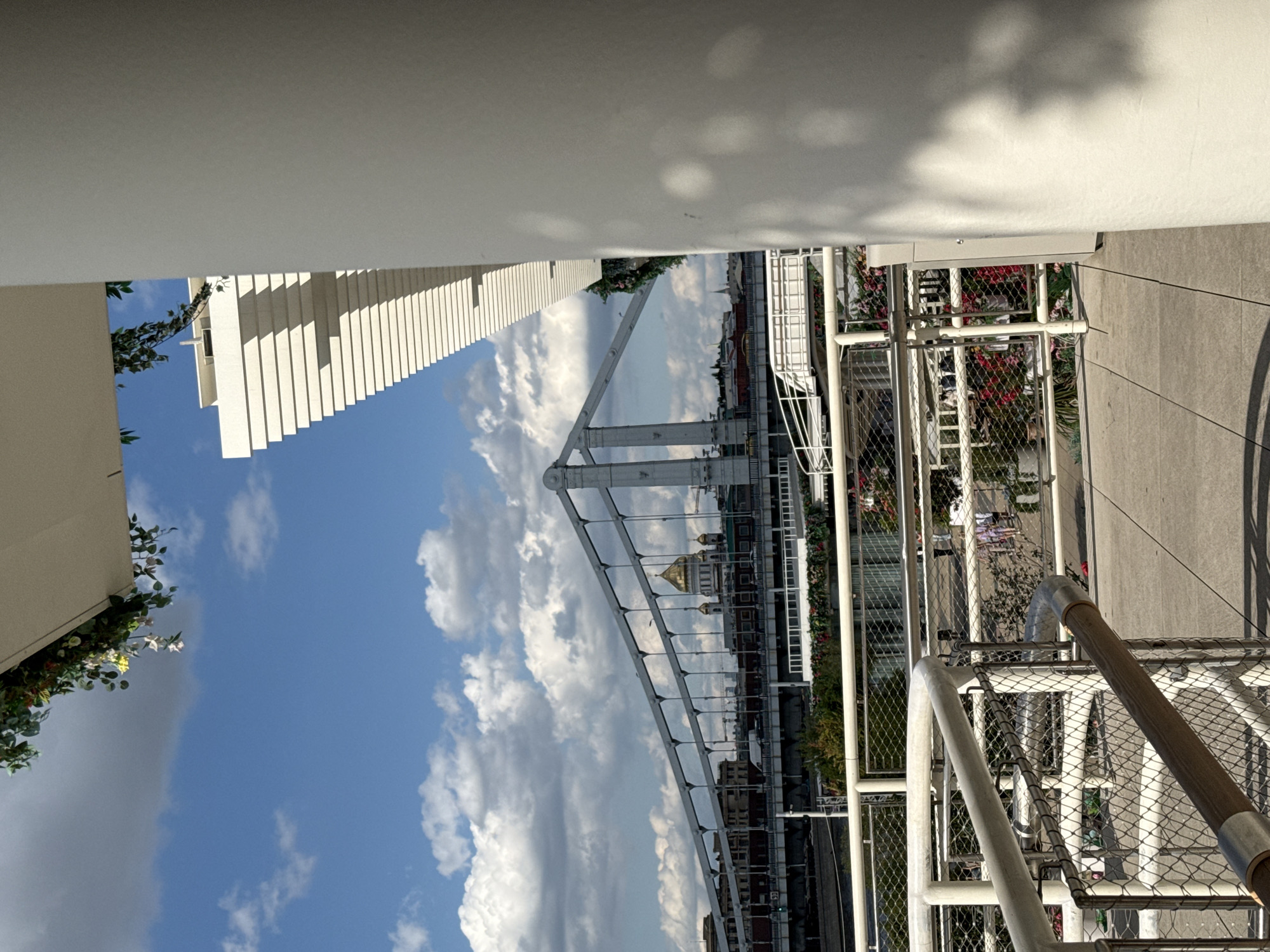 The image depicts a view from a balcony or terrace in Moscow, Russia, overlooking the Krymsky Bridge. In the foreground, a white railing with a wooden handrail encloses the outdoor seating area, which is paved with gray tiles. Beyond the railing, a white walkway with railings and some potted plants is visible.
The Krymsky Bridge, a suspension bridge, dominates the middle ground. Its distinctive white towers and cables span across the Moskva River. In the background, the golden domes of the Cathedral of Christ the Saviour are prominent, situated behind the bridge. Various buildings and trees line the riverbank on the other side.
The sky is blue with scattered white clouds. Lush green foliage with small flowers hangs from the overhang of the structure in the foreground, adding a touch of natural beauty to the urban landscape. The scene is bathed in sunlight, creating shadows on the terrace and the white pillar on the right side of the frame.