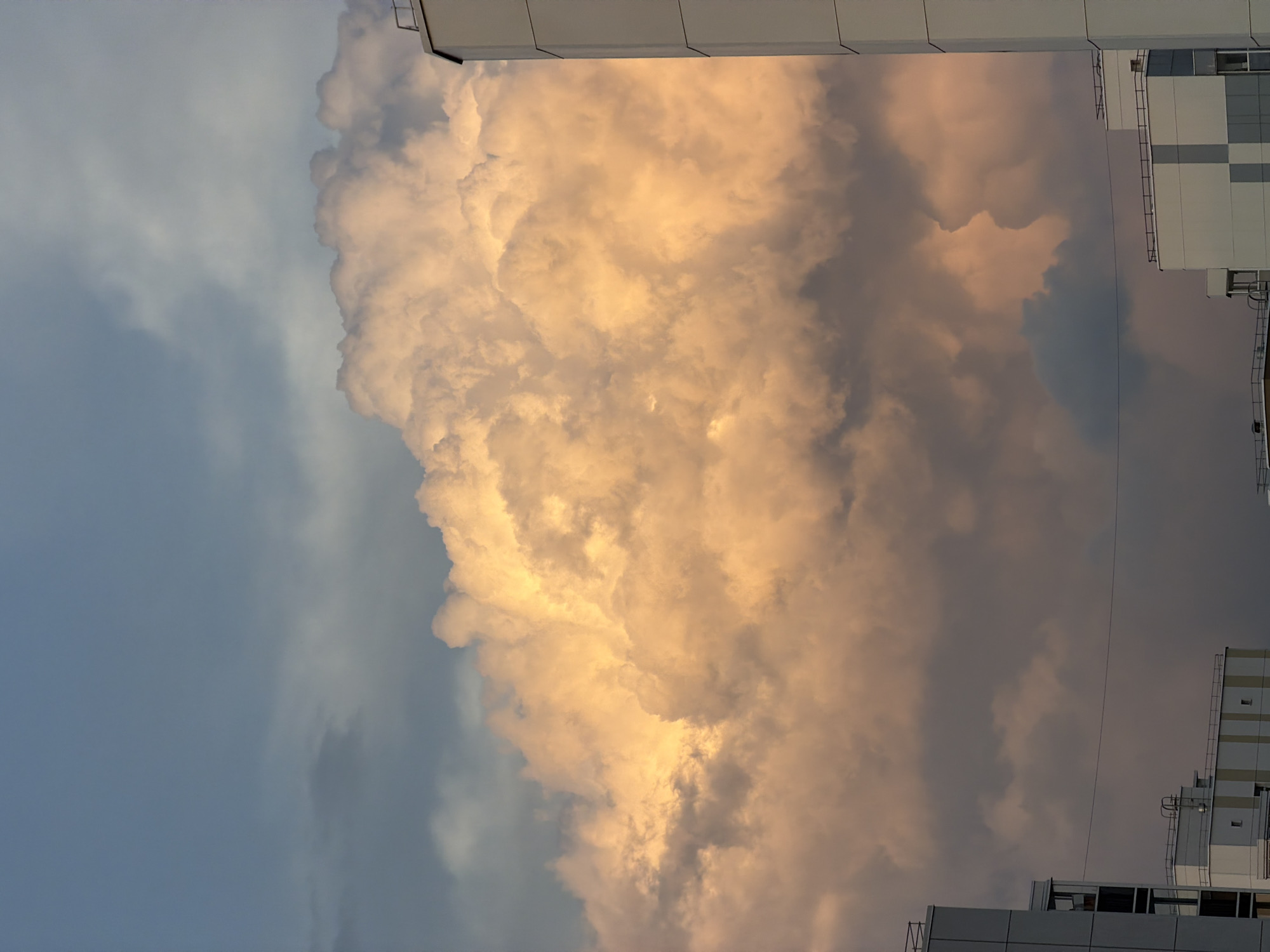 A wide-angle, eye-level photograph captures the Tsiolkovsky cityscape under a dramatic sky. A prominent, massive cumulus cloud dominates the central and upper portions of the frame, glowing with warm orange and yellow hues, suggesting it is illuminated by either sunrise or sunset. The cloud's texture is detailed, with fluffy, billowing formations and darker, shadowed areas. In the foreground, the upper sections of modern apartment buildings are visible on the right and left edges, their white and grey facades contrasting with the vibrant sky. A thin wire stretches horizontally across the mid-ground, intersecting the view of the buildings and clouds. The sky above the cloud is a gradient of pale blue and grey. No people are visible in the scene. The overall impression is one of urban architecture juxtaposed with a powerful natural sky phenomenon.