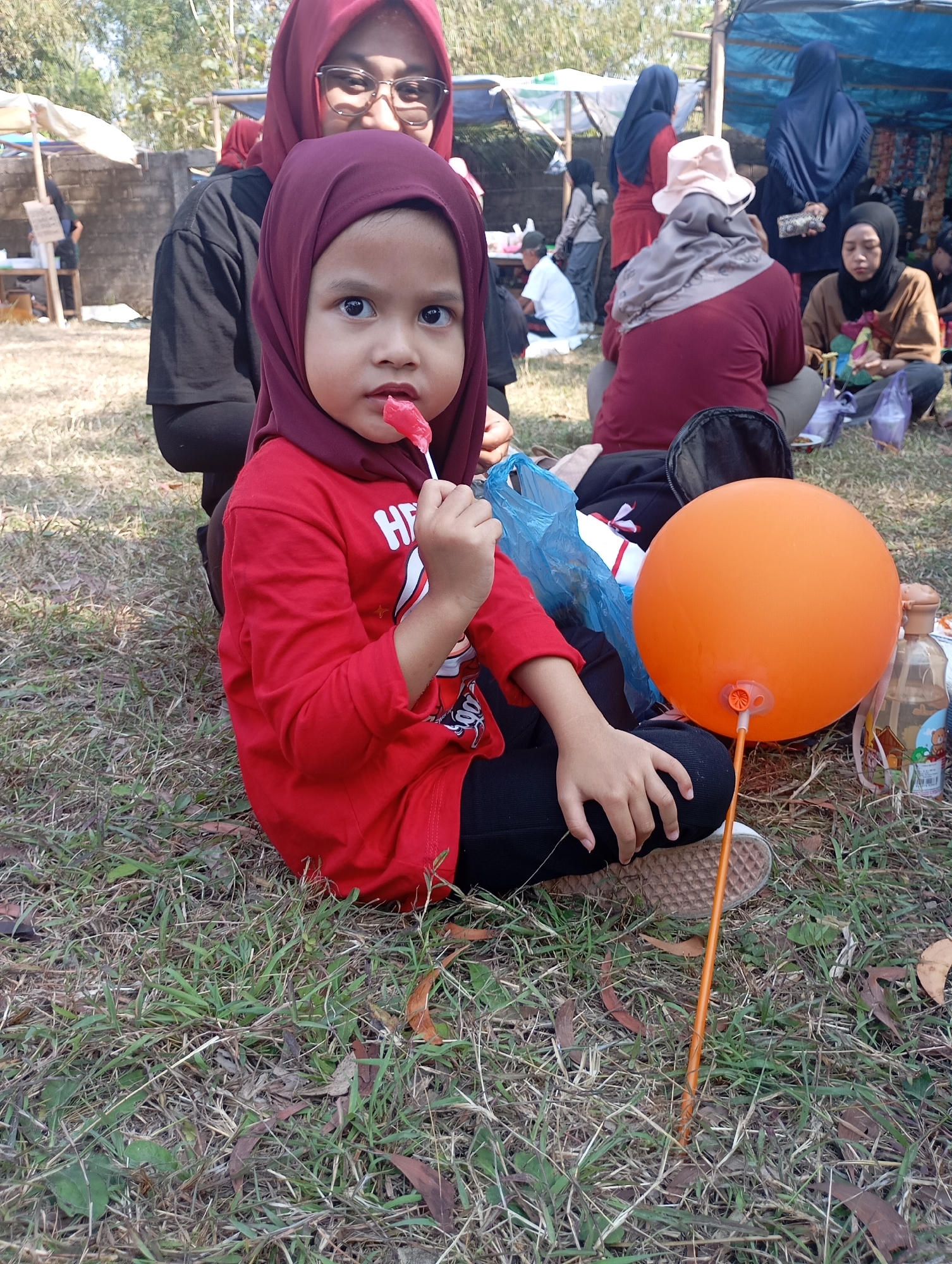 A young girl wearing a red hijab and a red long-sleeved shirt is sitting on the grass, holding a red lollipop. She is looking at the camera with wide eyes. Next to her, there is an orange balloon on a stick. In the background, several people are sitting and standing around, some wearing hijabs. The scene appears to be outdoors, possibly at a market or a gathering, with tents and trees visible. The ground is covered in dry grass and fallen leaves. The overall impression is of a casual outdoor event.