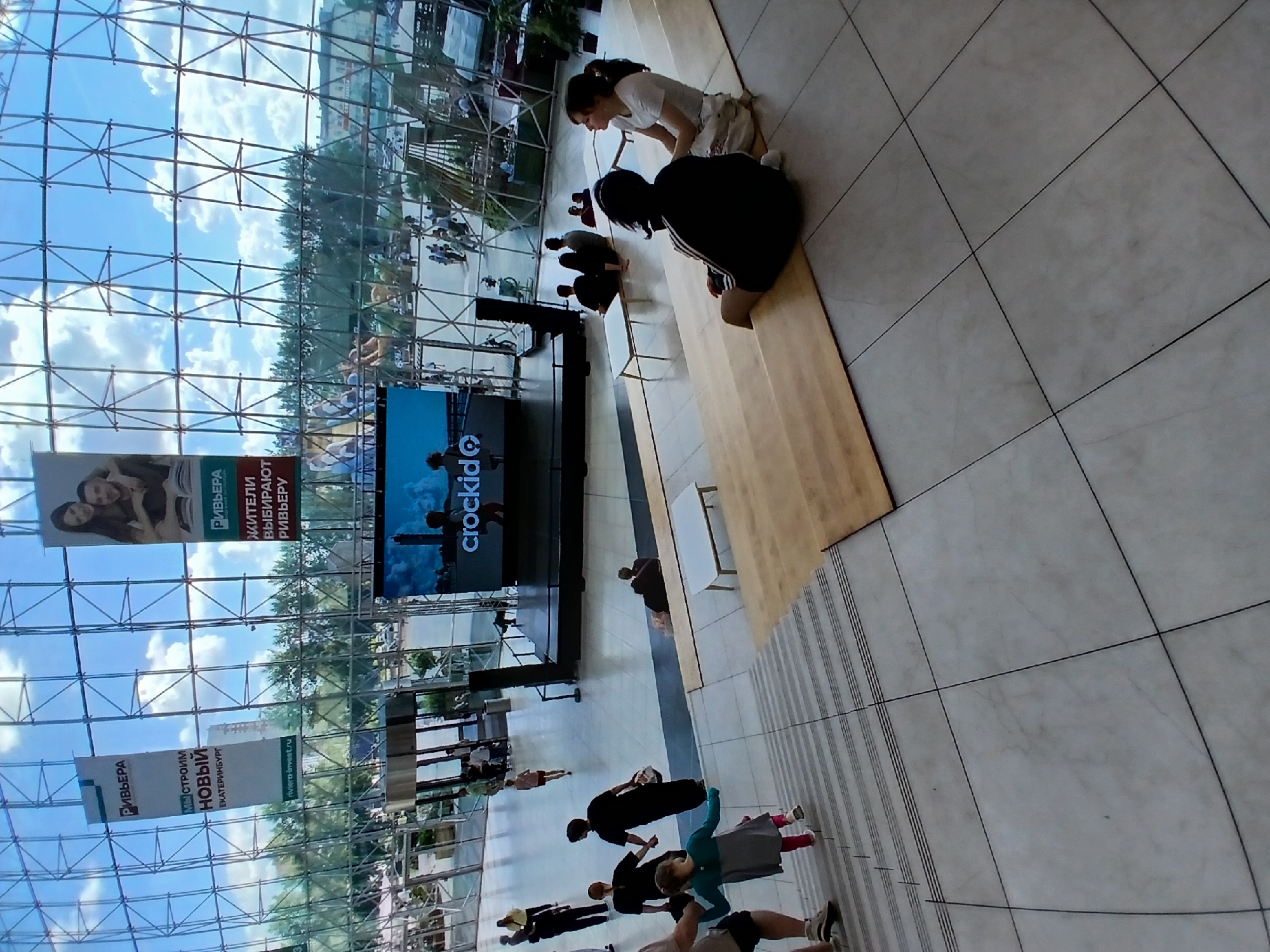 This image depicts the interior of a large, modern building, likely a shopping mall or exhibition center, with a distinctive geodesic dome-like glass roof. The scene is well-lit by natural light filtering through the glass and artificial lighting. In the foreground, a group of people are visible, including a man's legs and feet in shorts and sneakers, a young girl in a gray and teal dress walking up a wide staircase, and a woman in black holding a basket. Further up the stairs, another woman in a black top and pants is visible.
In the midground, there's a stage with a large screen displaying a logo that appears to be "crockid". Several people are seated on wooden tiered seating facing the stage. Two women are seated prominently on the right side, one in a white top and the other in a dark hooded jacket. Other individuals are scattered on the seating. Two small white tables are placed on the seating area.
In the background, through the glass roof, a cityscape is visible, including trees and buildings, suggesting an urban environment. There are also banners hanging from the structure, one of which has Russian text and the logo "РИВЬЕРА", which likely indicates "Riviera", and mentions "СТРОИМ НОВЫЙ ЕКАТЕРИНБУРГ" (We are building a new Yekaterinburg), placing the location in Yekaterinburg, Russia. Another banner shows a couple embracing. In the far background, more people are seen walking around outdoors, and some temporary structures or stalls might be present. The overall impression is a bustling public space during the daytime.