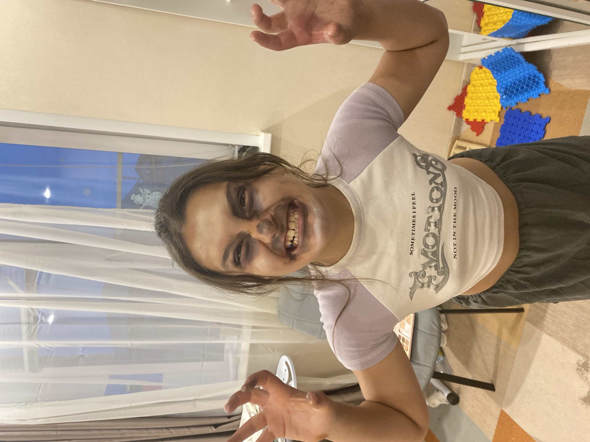 A female child or pre-teen is depicted from the waist up, centered in an indoor residential space in Tyumen, Russia, striking a menacing pose. She has dark brown hair and is wearing elaborate face makeup designed to appear ghoulish or zombified, featuring dark smudges around her eyes, a dark substance applied to her nose, and white makeup around her mouth and chin. Her mouth is open in a wide, snarling grin, baring her teeth, and her eyes are wide, conveying a frightful expression. Both hands are raised and splayed, fingers curled, also bearing dark smudges consistent with the face makeup. She is dressed in a white short-sleeved crop top with light purple raglan sleeves, featuring the text "SOMETIMES I FEEL EMOTION NOT IN THE MOOD," and grey athletic shorts.
The background includes a large window on the left with white sheer curtains, through which a bright blue sky and distant buildings are visible, indicating daytime. Snowflake decals and a snowman figure are affixed to the windowpane. To the left, a light grey upholstered chair and a small round table with a white top are partially visible. A cosmetic palette appears to be resting on the chair. The wall to the right is pale, and the floor features a multi-colored pattern of beige, orange, and white. Several interlocking plastic play mats in blue, yellow, and red are scattered on the floor in the bottom right corner, partially reflected in a vertical mirror or reflective surface.