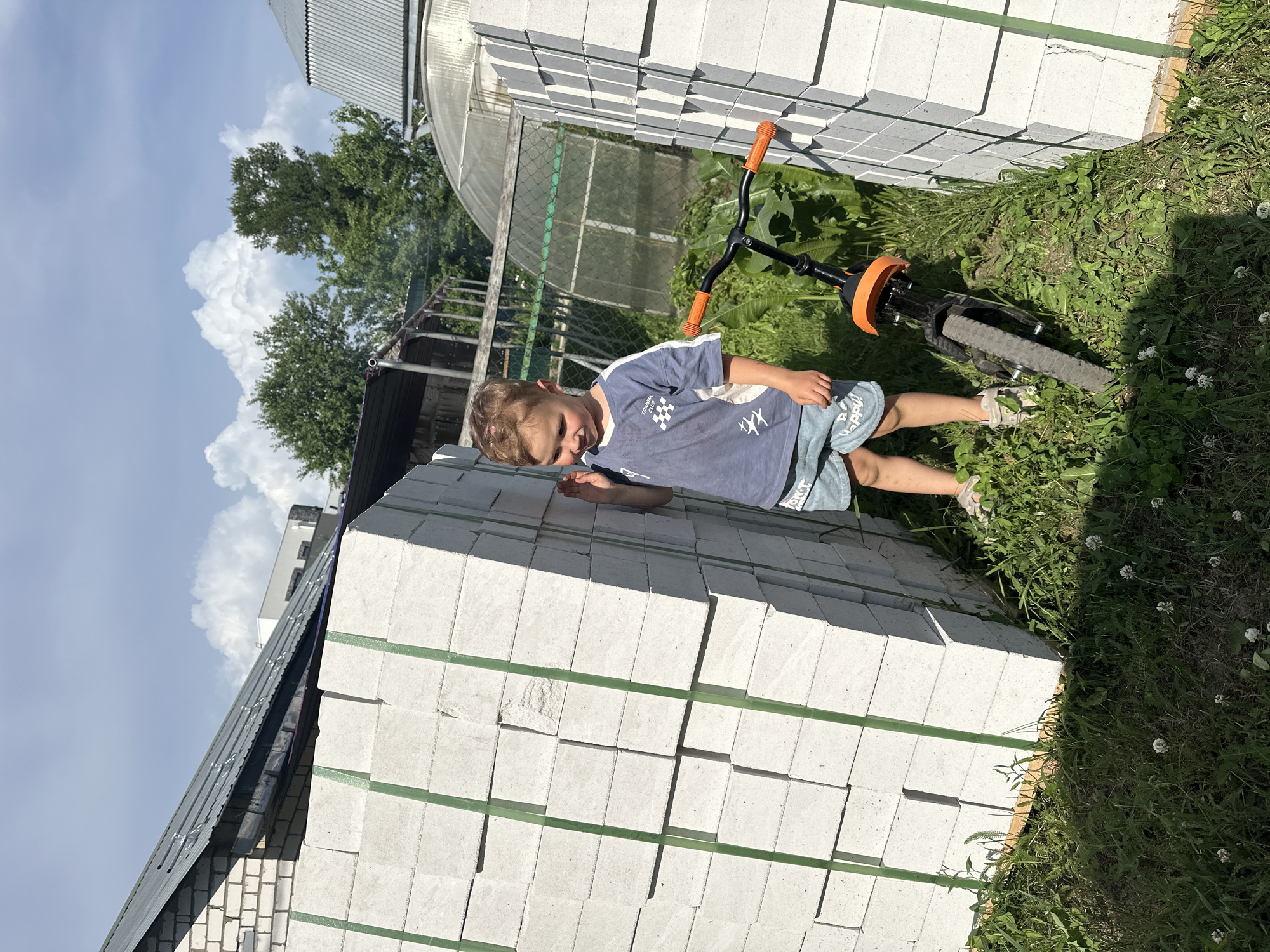 The image shows a young boy standing outdoors next to a large stack of white building blocks. He is wearing a blue t-shirt with a graphic design and blue shorts. A black and orange bicycle is parked beside him, leaning against the blocks. In the background, there is a greenhouse, trees, and parts of buildings with metal roofs. The scene appears to be in a residential or construction area. The location is specified as Nizhny Novgorod, Russia.