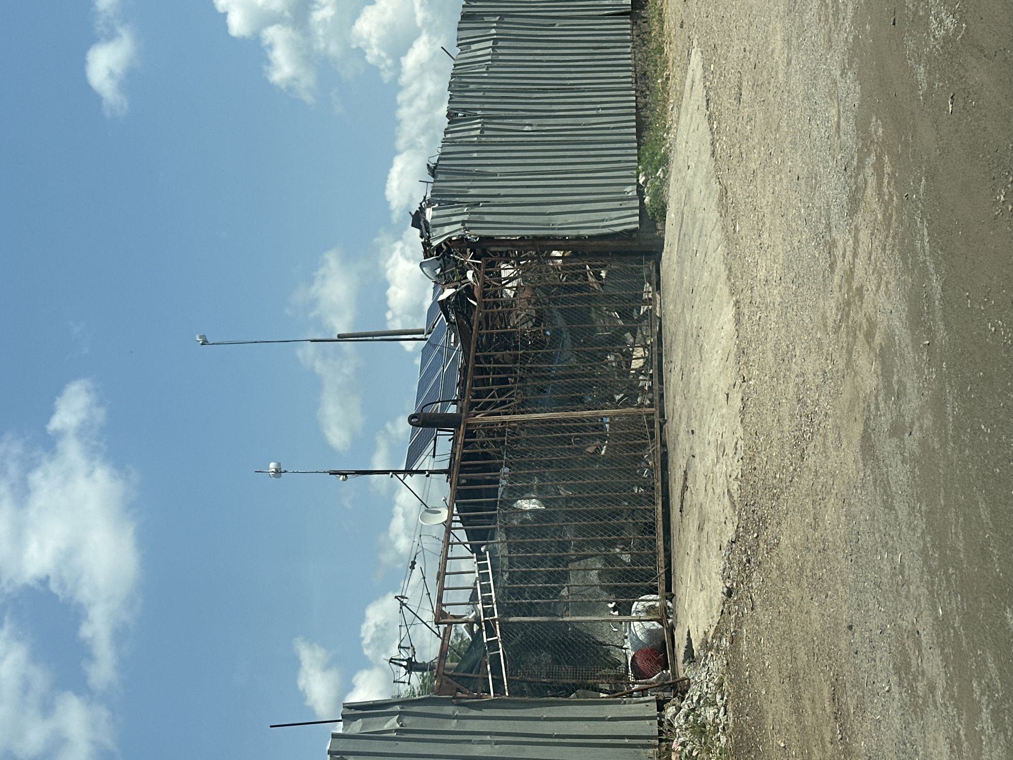 The image displays an outdoor scene under a clear blue sky with scattered white clouds, captured in daylight. The primary focus is a fenced-off enclosure, likely a storage area or junkyard, constructed with corrugated metal sheets forming walls and a rusty metal grate acting as a gate or part of the fence structure.
Inside the fenced area, various objects are visible. A large solar panel is prominently placed on top of the structure, angled towards the sky. Below it, behind the metal grate, are numerous indistinct items, appearing as scrap metal, discarded machinery, or covered bulk objects, possibly under netting or tarp. A metal ladder leans against the fence on the left side, and a small satellite dish is also present within the enclosure. Near the bottom right of the fenced area, a white and red container or bag is discernible among debris.
Along the upper edge of the image, extending into the blue sky, are two slender poles, possibly street lights or utility poles, each topped with a small, light-colored device that could be a camera or sensor.
To the right of the fenced enclosure is an unpaved, light-colored dirt or gravel road or path. The ground shows signs of wear and loose stones. No people are visible, and no specific activity is taking place. There is no visible text. The overall impression is that of an utilitarian or informal outdoor storage area in an industrial or semi-urban setting.