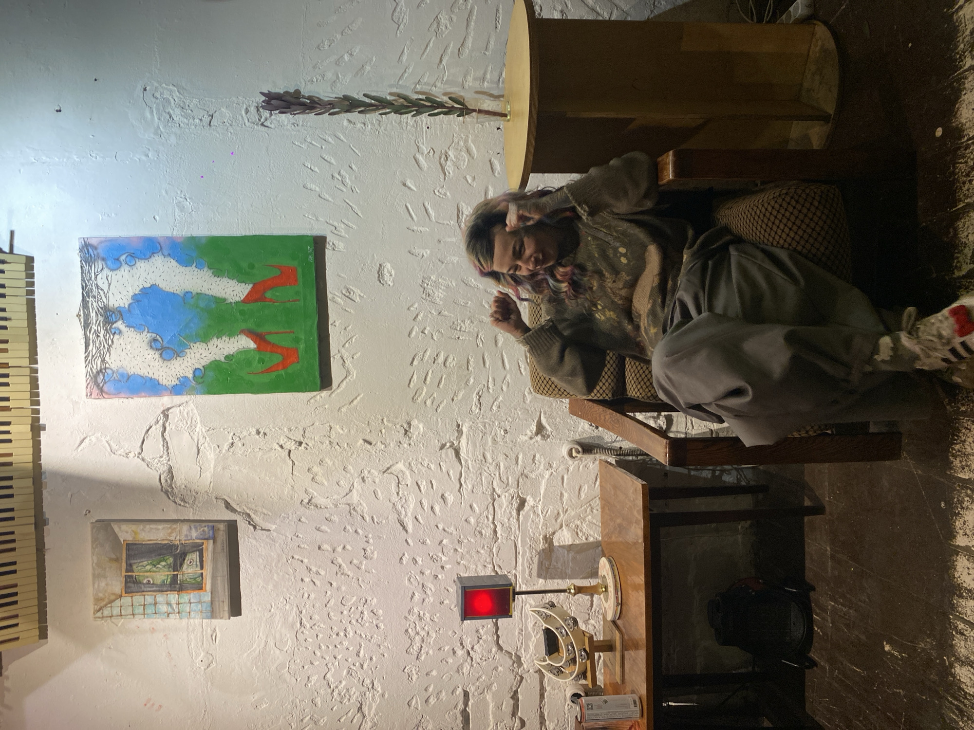 A person sits in a chair next to a table in an interior space with textured white walls. The person has dyed hair and is wearing a patterned sweater and gray pants. On the wall are several items, including what appears to be keys from a piano or keyboard, two paintings, and an object with a red light. The location cannot be definitively identified as Moscow, Russia, based on the visual information provided.