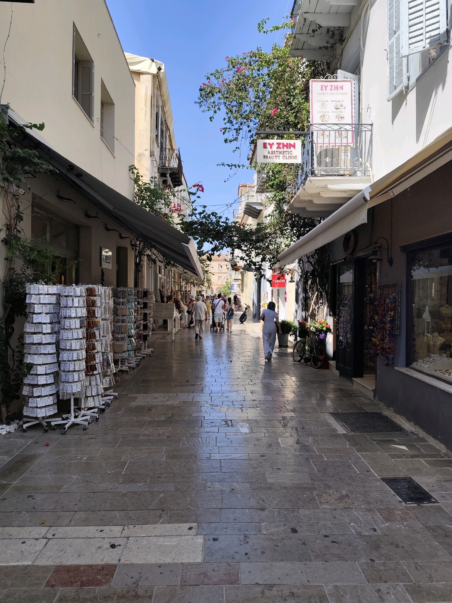 This image depicts a sun-drenched pedestrian street in Nafplio, Greece. The street is paved with large, uneven stone slabs, some of which appear wet and reflective. Buildings with pale facades line both sides of the street, featuring awnings and balconies.
On the left side, a shop displays rows of small items on stands, likely souvenirs or trinkets. Further down, several people are browsing or walking. Above this shop, a balcony has a sign for "EY ZHN AESTHETIC BEAUTY CLINIC."
On the right side, more shops are visible, with their doorways and windows open. A bicycle adorned with flowers is parked in front of one establishment. A woman in a light-colored outfit walks away from the camera down the street. More people are seen further in the distance. Overhead, trees with pink and green foliage create dappled shade. The sky is a clear, bright blue. The overall impression is a lively, picturesque street scene in a European town.