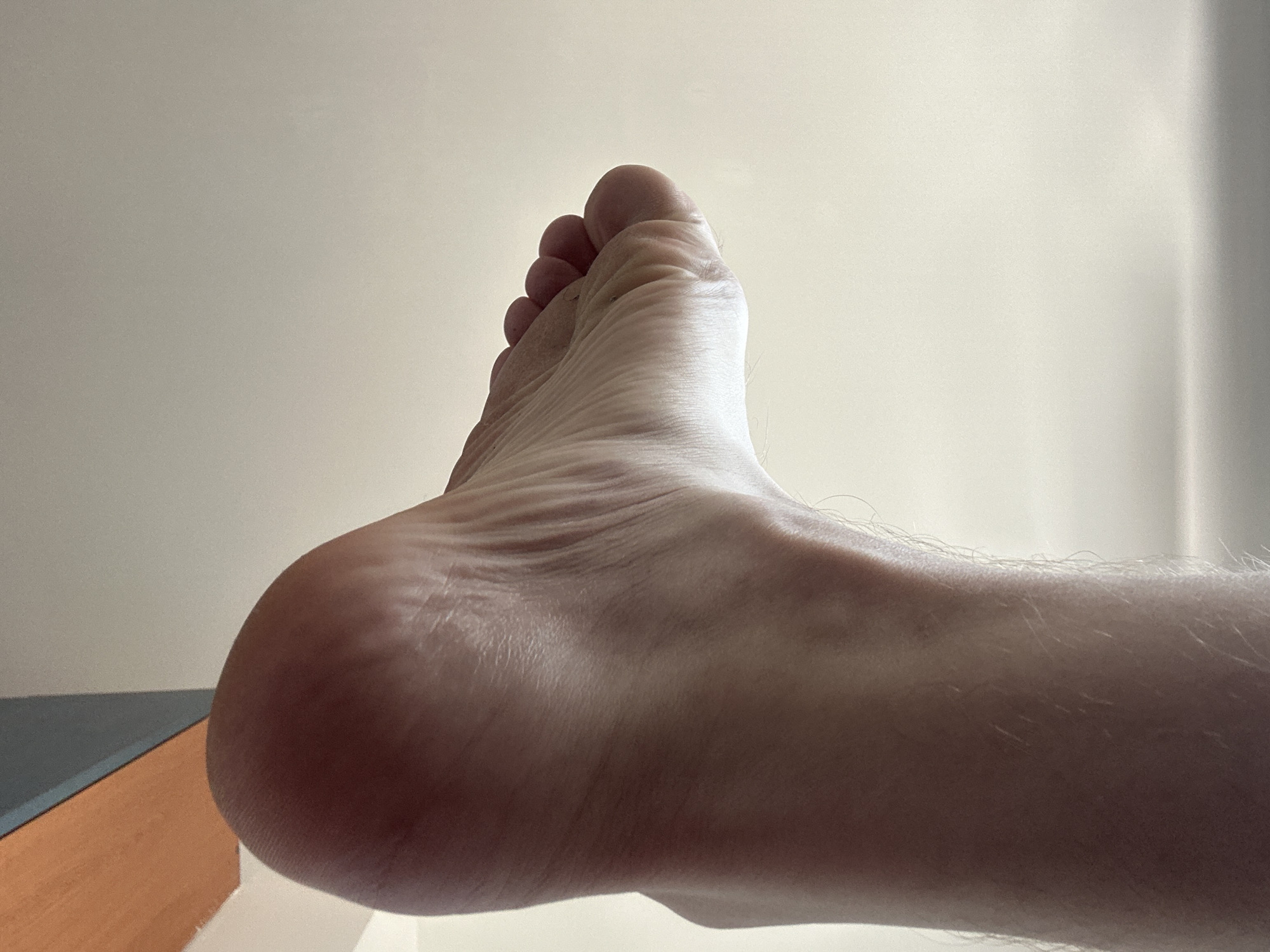 An extreme close-up, low-angle shot shows the sole and arch of a bare human foot, positioned as if stepping down from a surface. The skin is pale with visible texture and fine hairs on the lower leg. The foot is illuminated by a light source from the right, casting subtle shadows that highlight the contours of the arch and toes. In the upper left corner, a small section of an orange and gray striped surface is visible, suggesting the foot is interacting with an elevated platform or furniture. The background is a plain, light-colored surface, possibly a wall or ceiling. The image does not contain any information that can be used to determine its location in Zvezdara Urban Municipality, Serbia.
