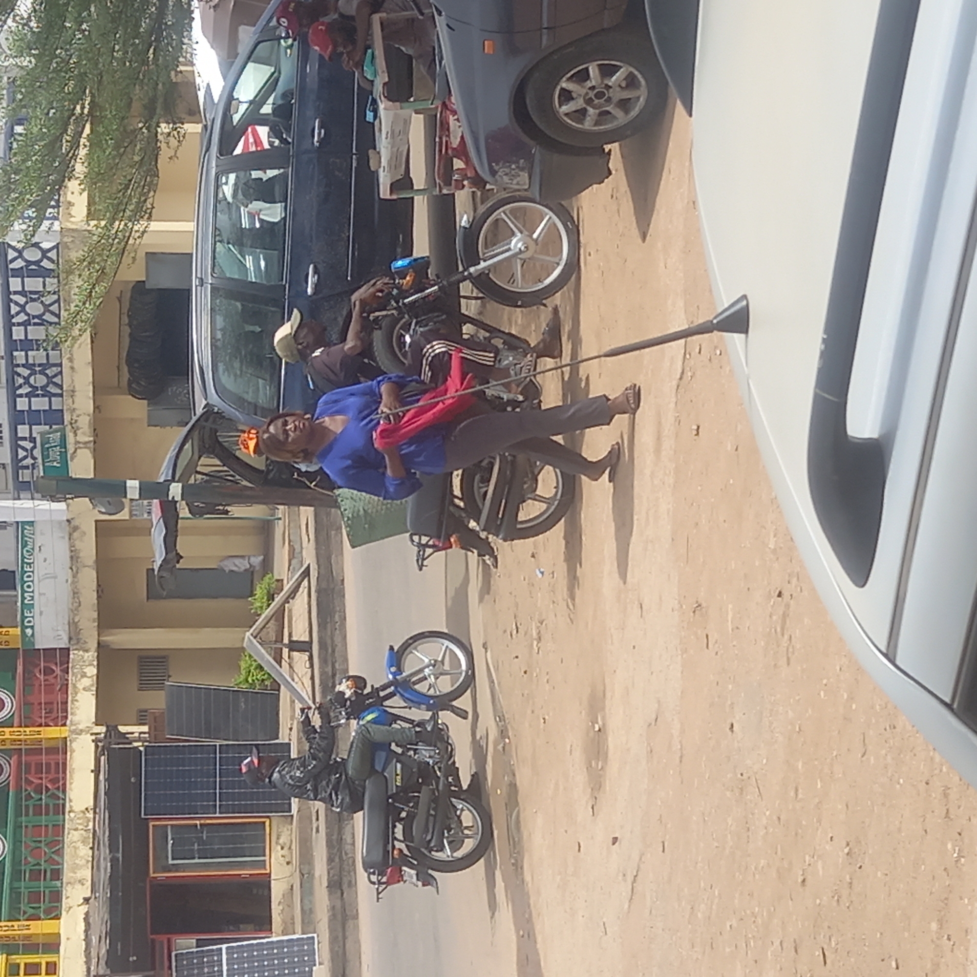The image depicts a street scene in Dadi Riba, Nigeria. In the foreground, a white vehicle is parked. Adjacent to it, a woman in a blue shirt and gray pants is standing by a black motorcycle. She is holding a red bag. Another man wearing a beige cap is on a similar motorcycle in front of a dark minivan, on which are a few people with red hats. In the background, solar panels are leaning against a building wall. A pole is going from a motorcycle to the white car.