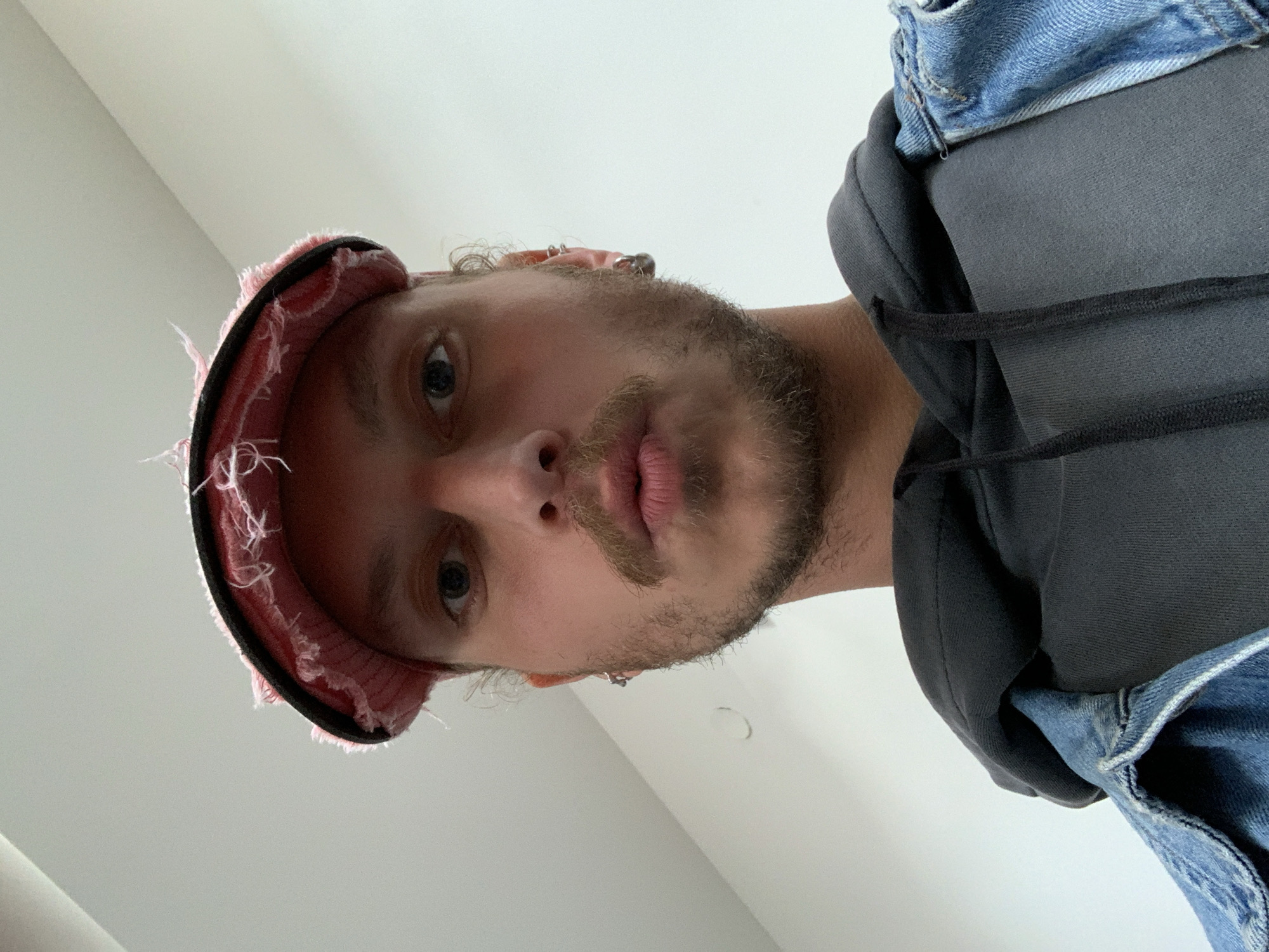 The image presents a close-up, low-angle portrait of a single male individual. The subject has light-colored eyes, full lips slightly pursed, a mustache, and a beard covering the jawline and chin. He wears a distressed pink-red flat cap with a black brim, a dark grey hooded sweatshirt with the hood up, and a light blue denim jacket over it. Multiple silver-toned hoop earrings are visible in both ears. The background is a plain, uniformly light grey wall, indicative of an indoor setting. There are no specific environmental details visible within the image to identify a precise location in the city of Tel-Aviv, Israel, or any other city. The individual is directly facing the camera.