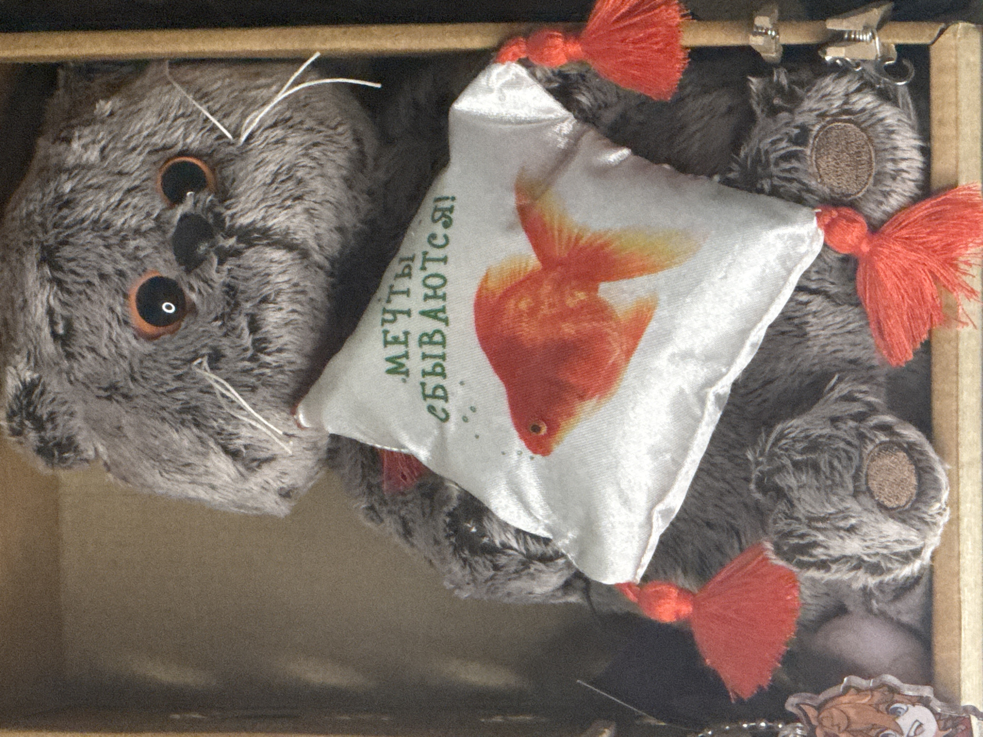The image shows a plush gray cat toy with orange-brown eyes, white whiskers, and a black nose, sitting inside a wooden box. The cat is holding a white square pillow embroidered with a golden fish and green Cyrillic text that reads "Мечты сбываются!" (Dreams come true!). Red tassels are attached to the corners of the pillow and also on the cat's paws. To the left of the cat, a keychain with a character illustration is visible. The scene is indoors, likely a shelf or display within a home. There is no information in the image to suggest its location in Saratov, Russia.