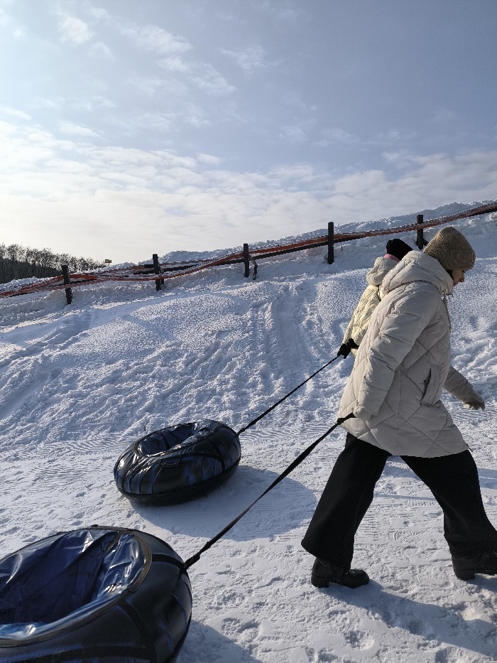 The image captures a winter scene on a snow-covered slope in Prybuzke, Ukraine, under a partially cloudy blue sky. A person, dressed in a light-colored puffer coat, dark trousers, and a light-colored beanie, walks from right to left across the frame, pulling two dark, patterned inflatable snow tubes uphill by black straps. A second person, partially visible behind the first, wearing a light-colored puffer jacket and a dark hat, is also ascending the slope. The slope exhibits visible tracks in the textured snow. In the background, a fence composed of dark posts and red-orange horizontal banding runs along the crest of the hill. A line of dark trees is visible on the distant horizon.