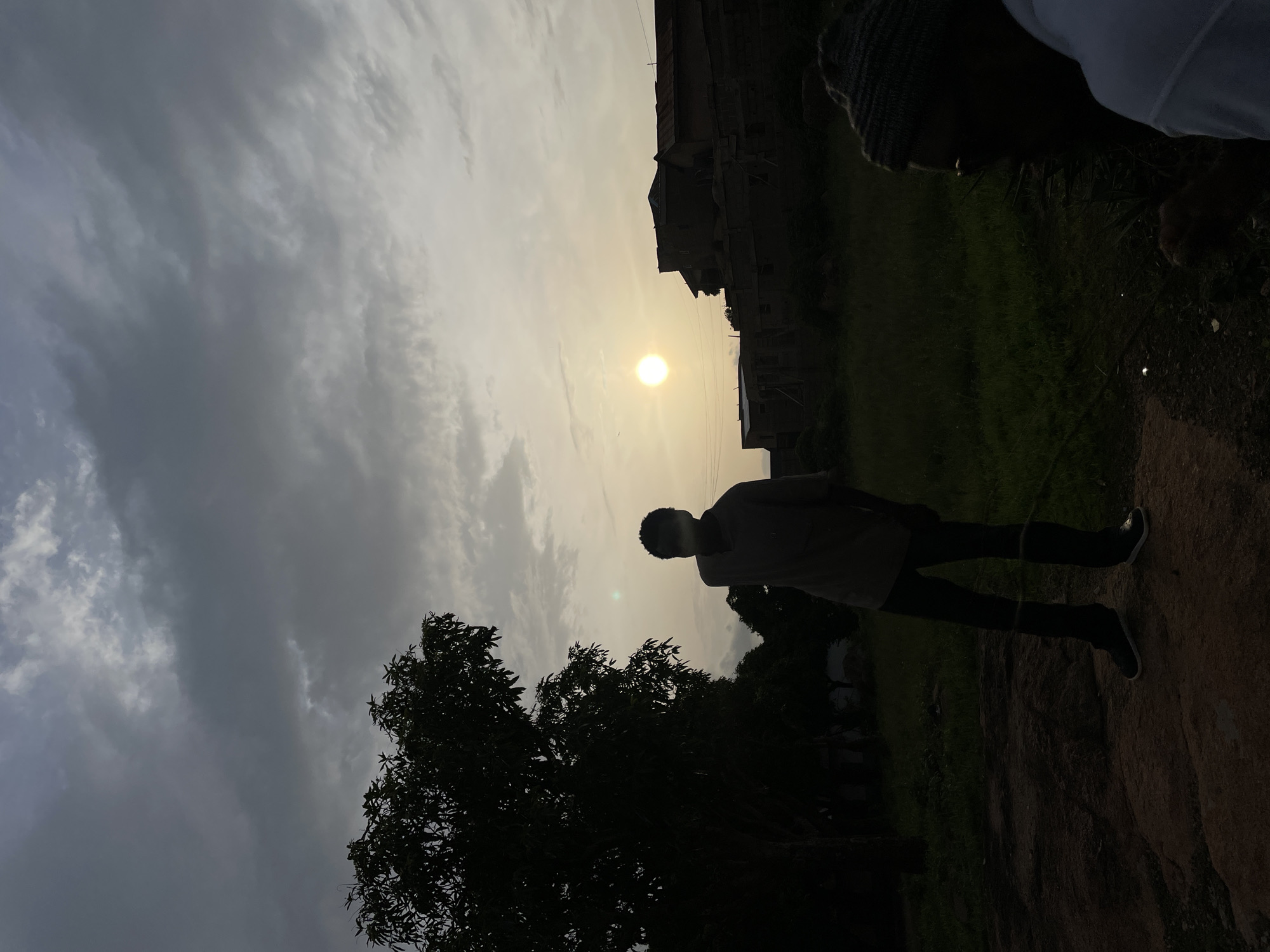 The image shows an outdoor scene in Jos, Nigeria, at dusk or dawn. The sky is filled with grey, textured clouds, and the sun is a bright disc. Silhouetted against the sky is a person standing on a rocky or earthen surface. They are wearing dark pants, a light-colored shirt, and shoes. A tree with dense foliage is silhouetted in the lower left of the frame. In the background, a building is visible, also silhouetted. Another person partially enters the frame from the top right. The focus is on the figure standing silhouetted against the sun.