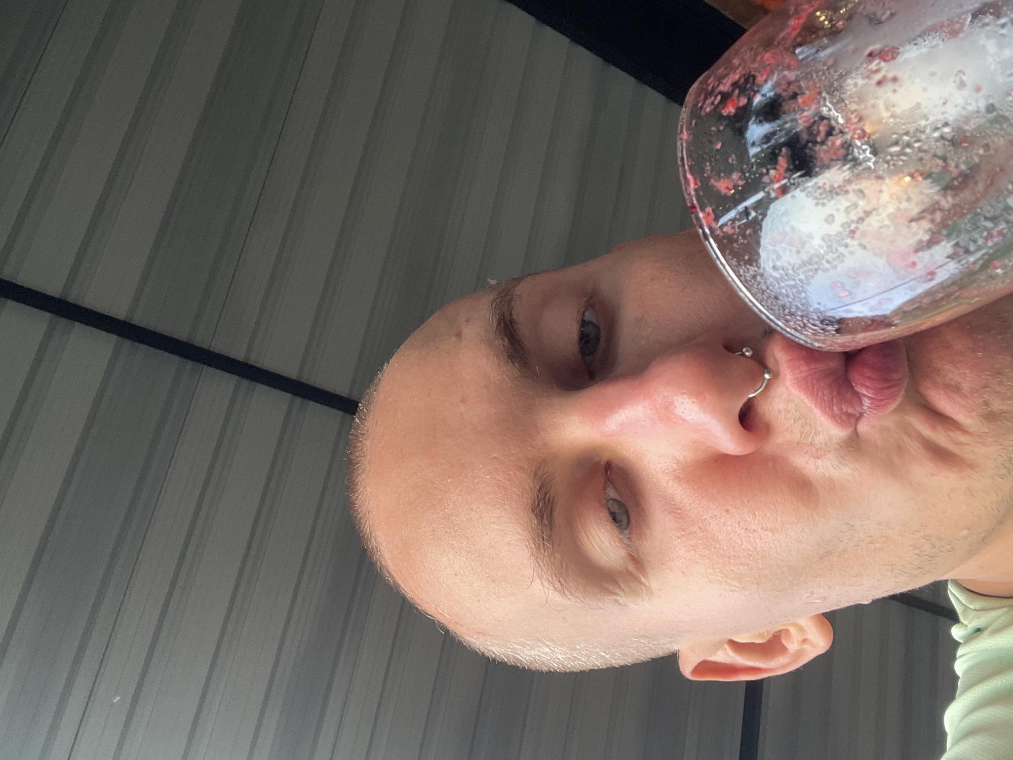 A close-up, low-angle shot depicts a person drinking from a glass. The person appears to be bald with fair skin, light-colored eyes, and a nose ring. The glass is tilted toward the viewer, revealing remnants of a red-colored beverage. The background consists of corrugated metal panels. There is no definitive information to suggest the location is Tambon Nam Phrae, Thailand.