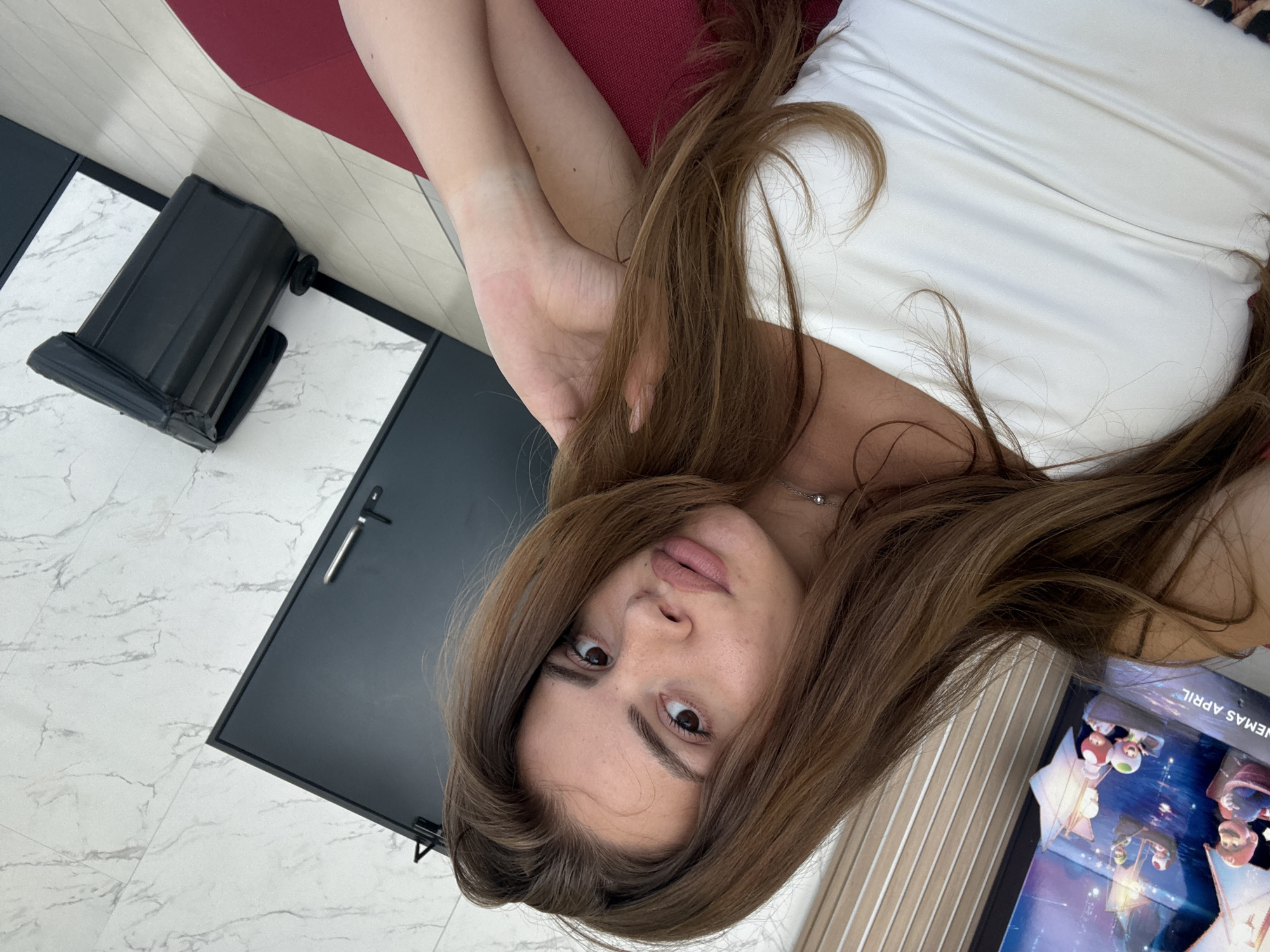 A young woman with long brown hair and a white tank top is sitting down, posing for a selfie. She is indoors, possibly in a public space with a movie poster in the background. The poster appears to be for "The Super Mario Bros. Movie," which was released in April. The environment is modern, with marble-patterned walls and light-colored flooring. A black trash bin is visible in the background, near a door. There is no indication this scene is in Plovdiv, Bulgaria.