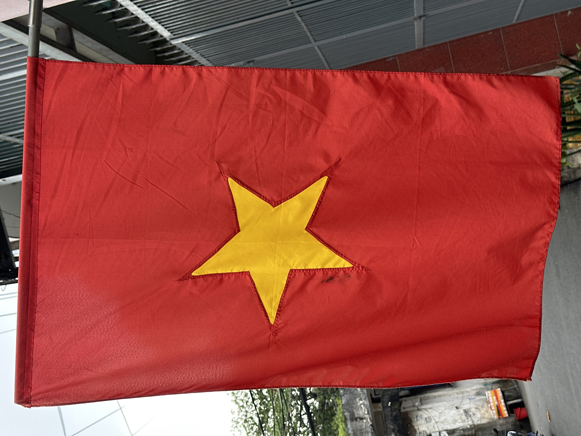 A close-up shot reveals a Vietnam flag, its red fabric marked with wrinkles and creases, suspended outdoors. A large, centrally placed yellow star occupies the flag's foreground. Hints of the environment suggest an urban setting with glimpses of buildings, greenery, and possibly city infrastructure visible in the background. Given the urban setting and the presence of the Vietnam flag, the image is likely taken in a populated area of Đồng Nai, Vietnam, though a specific location within the city cannot be determined from the given image.
