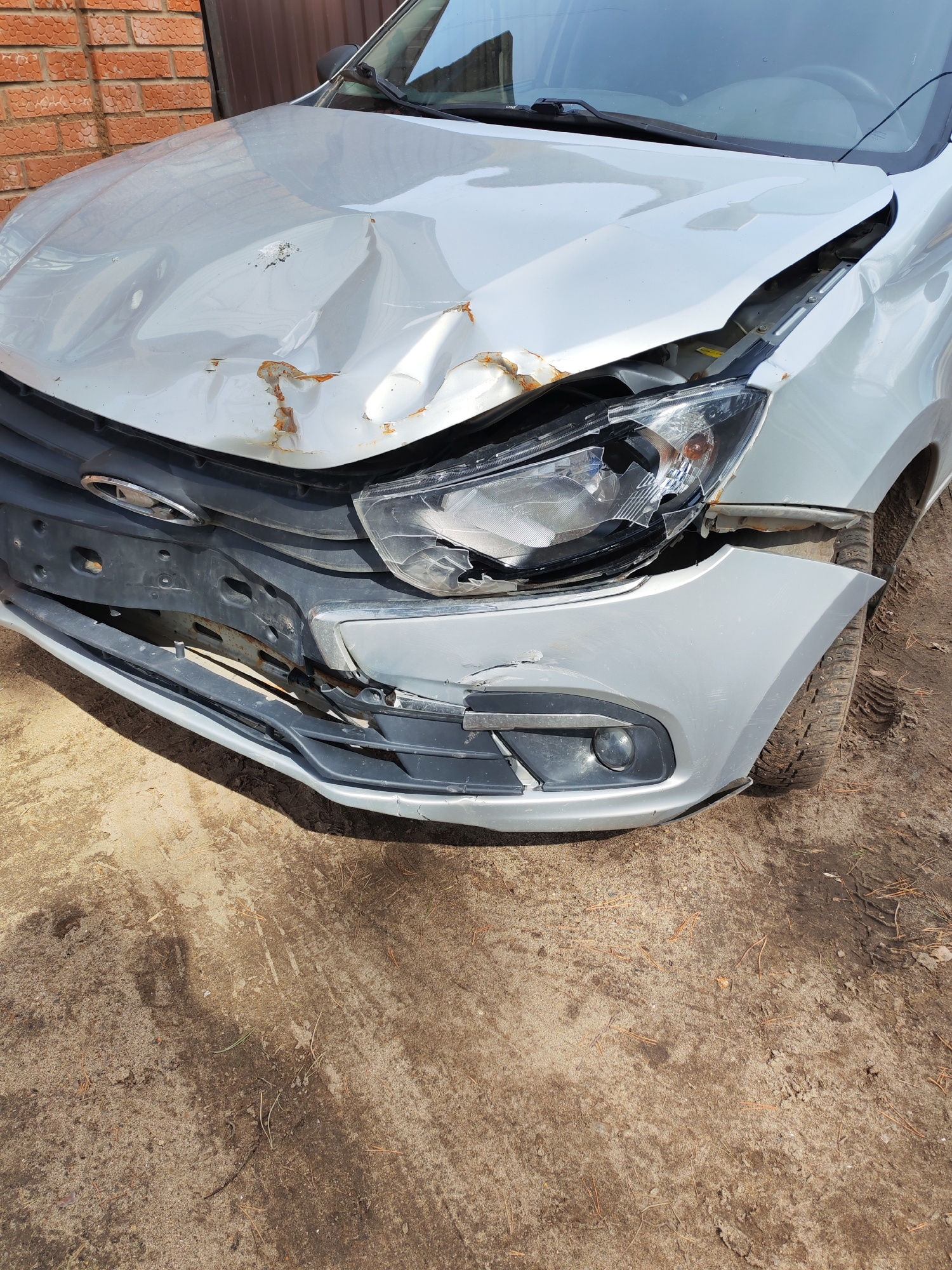 The image depicts the front end of a silver car with significant damage, likely from a collision. The hood is crumpled, and the front passenger-side headlight is shattered. The bumper is also damaged, with pieces broken off or displaced. Some rust is visible on the damaged metal. The car is situated on a dirt and sand surface. The scene is near a brick wall of a building. The location is presumed to be in the city of Chaikovsky, Russia. There are no people visible in the image.