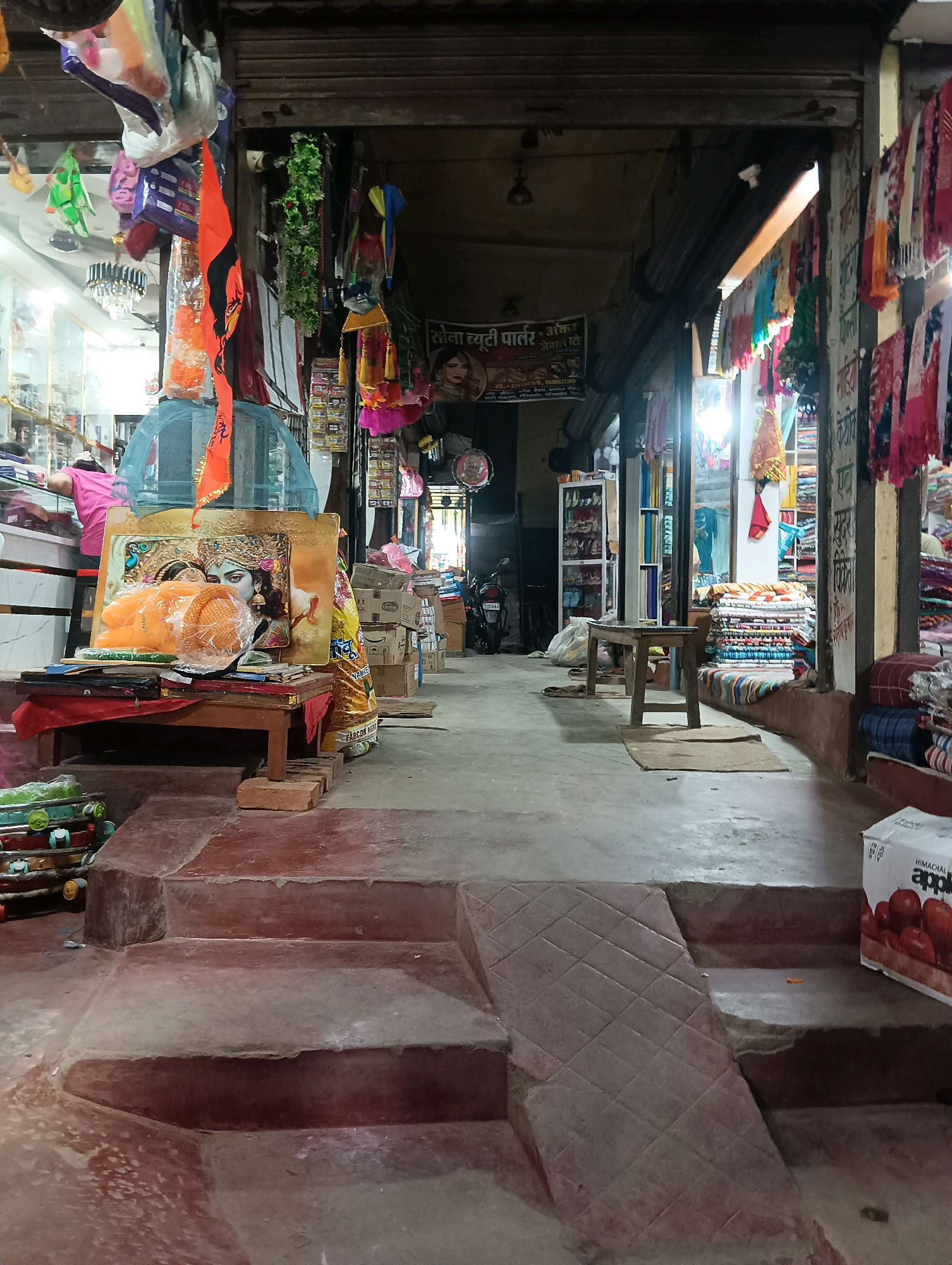 The image shows the entrance to a market in Dakhin Parsa, India. The scene is a busy street with various shops and stalls. In the foreground, there are steps leading up to the market. The steps are made of concrete and have a reddish-brown color. To the left of the steps, there is a stack of colorful items, possibly toys or goods.
The market entrance is a wide opening with various items displayed. On the left side, there are shelves stocked with goods, and a person in a pink shirt is visible behind a counter. In the center, a religious picture of a deity, possibly Krishna, is displayed on a wooden stand. The picture is framed in gold and has orange fabric draped around it. There are also decorative items hanging from the ceiling, including colorful flags and garlands.
To the right of the entrance, more stalls are visible, with stacks of colorful fabrics and other merchandise. A wooden bench is placed in front of one of the stalls. The overall environment appears to be a typical Indian market, with a lot of activity and a vibrant atmosphere. The lighting is dim in some areas and brighter in others, suggesting a mix of indoor and outdoor spaces.