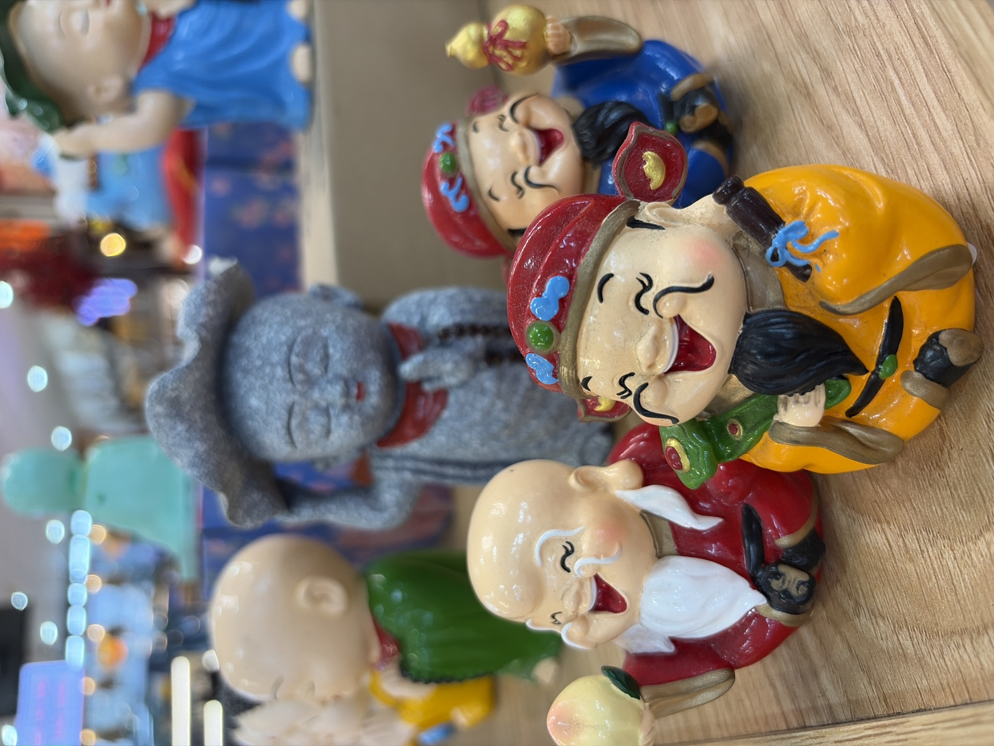 A group of small, smiling, and cheerful ceramic figurines are arranged on a wooden surface. The figurines depict plump figures with round faces, closed eyes, and wide smiles, suggesting joy or good fortune. One figurine, in the foreground and to the left, wears a red robe with white trim and holds a light-colored peach in its left hand. It has a long white beard and a small green frog on its chest. To its right, another figurine wears a bright orange robe and holds a dark object with a blue ribbon tied around it in its right hand. This figure also has a black beard and a green element on its chest. In the background, a grey stone-like figurine with a hat appears to be gesturing. Further back, a light-skinned figurine in a green robe is visible, and behind that, a figurine in a blue robe with a green hat. The overall impression is of a display of decorative cultural figures. The scene is set indoors with artificial lighting, and the wooden surface suggests a shelf or display stand. The blurred background indicates other items or displays are present but out of focus. There is no indication of the city of Da Nang, Vietnam, or any specific city.