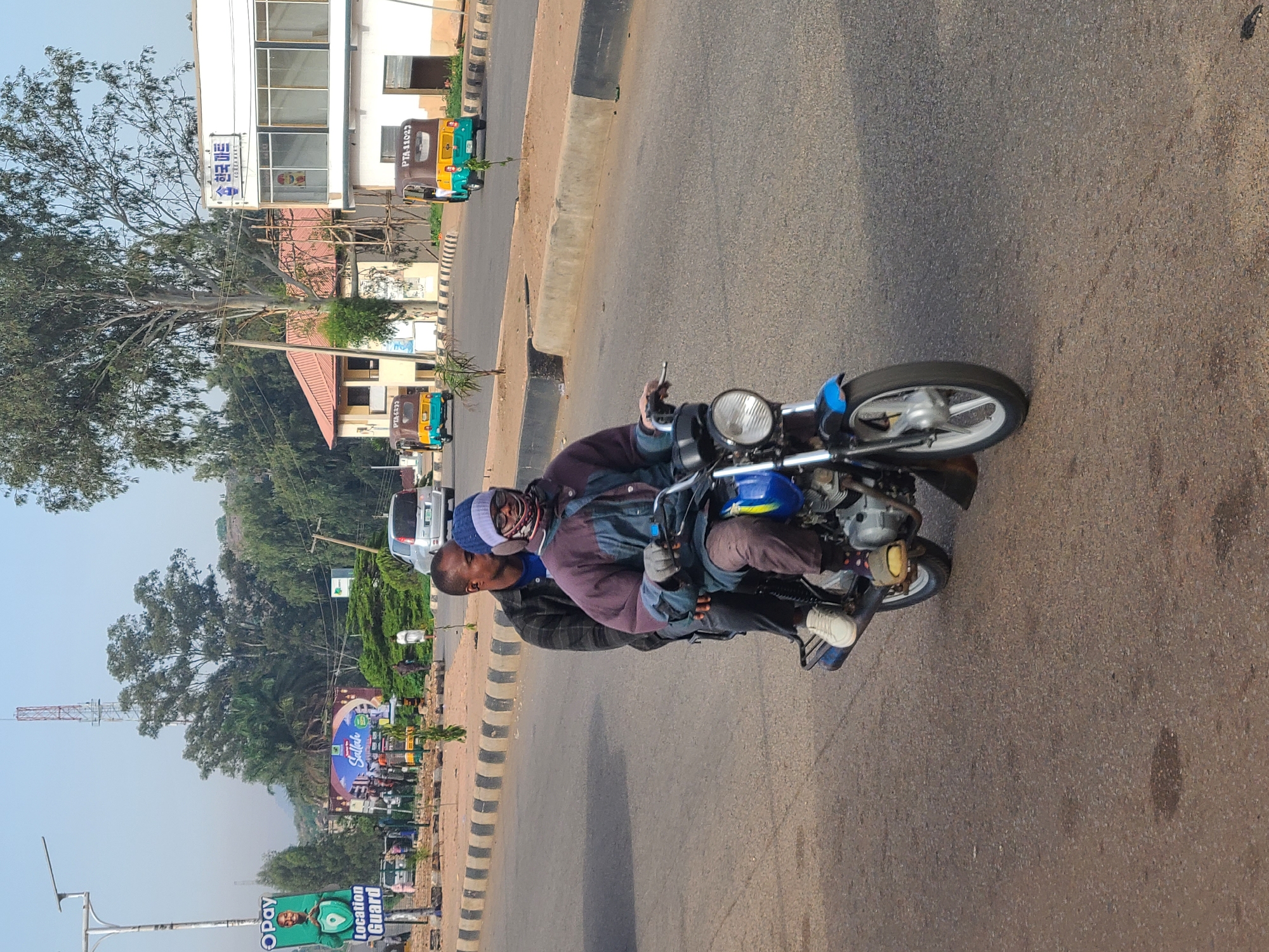 A motorcycle with two riders is traveling along a road in Gura Top, Nigeria. The rider in front is holding a cell phone in his left hand. The other rider is behind him. The road is bordered by buildings, trees, and billboards on either side. The area appears to be a commercial or urban district. The sky is clear.
