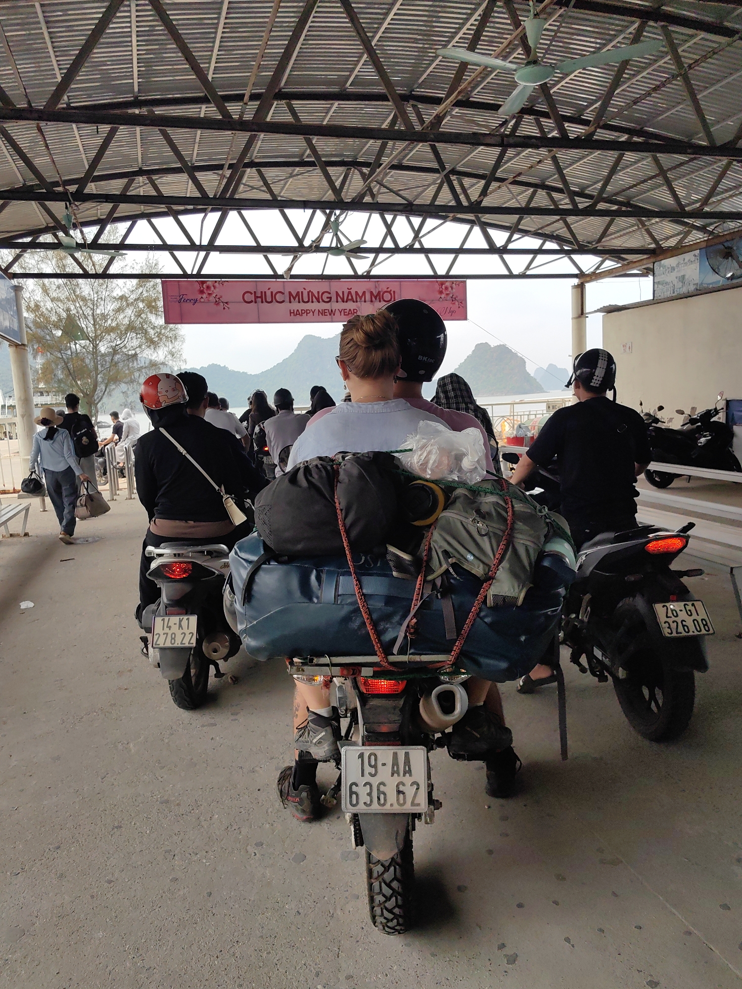 The image shows a group of people on motorcycles, presumably tourists, waiting in line at a ferry terminal. The terminal is located in the city of Hải Phòng, Vietnam. The motorcycles are loaded with luggage, indicating that the travelers are likely on a long journey. In the background, mountains can be seen through the ferry terminal's open entrance. Above the line of motorcyclists, there is a sign with the text "CHÚC MỪNG NĂM MỚI" (Happy New Year) printed on it. The license plates of the motorcycles read "14-K1 278.22", "19-AA 636.62" and "26-G1 326.08".