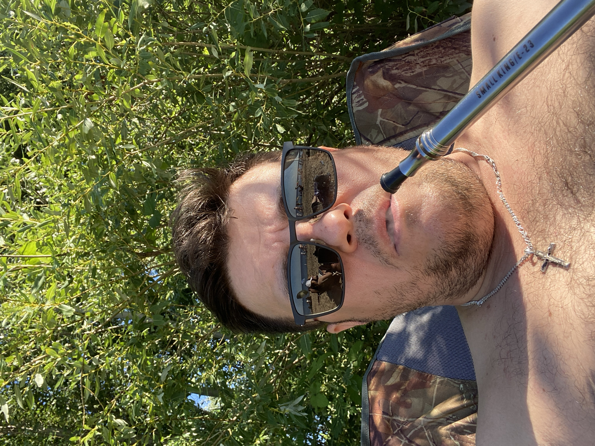 In Novi Petrivtsi, Ukraine, a man sits outdoors, partially visible in a close-up shot, using a vape pen. He is wearing sunglasses that reflect a beach scene with people and umbrellas. The man is shirtless, adorned with a silver chain and a cross pendant. He is seated in a chair with a camouflage pattern. Lush green foliage forms the background of the image.