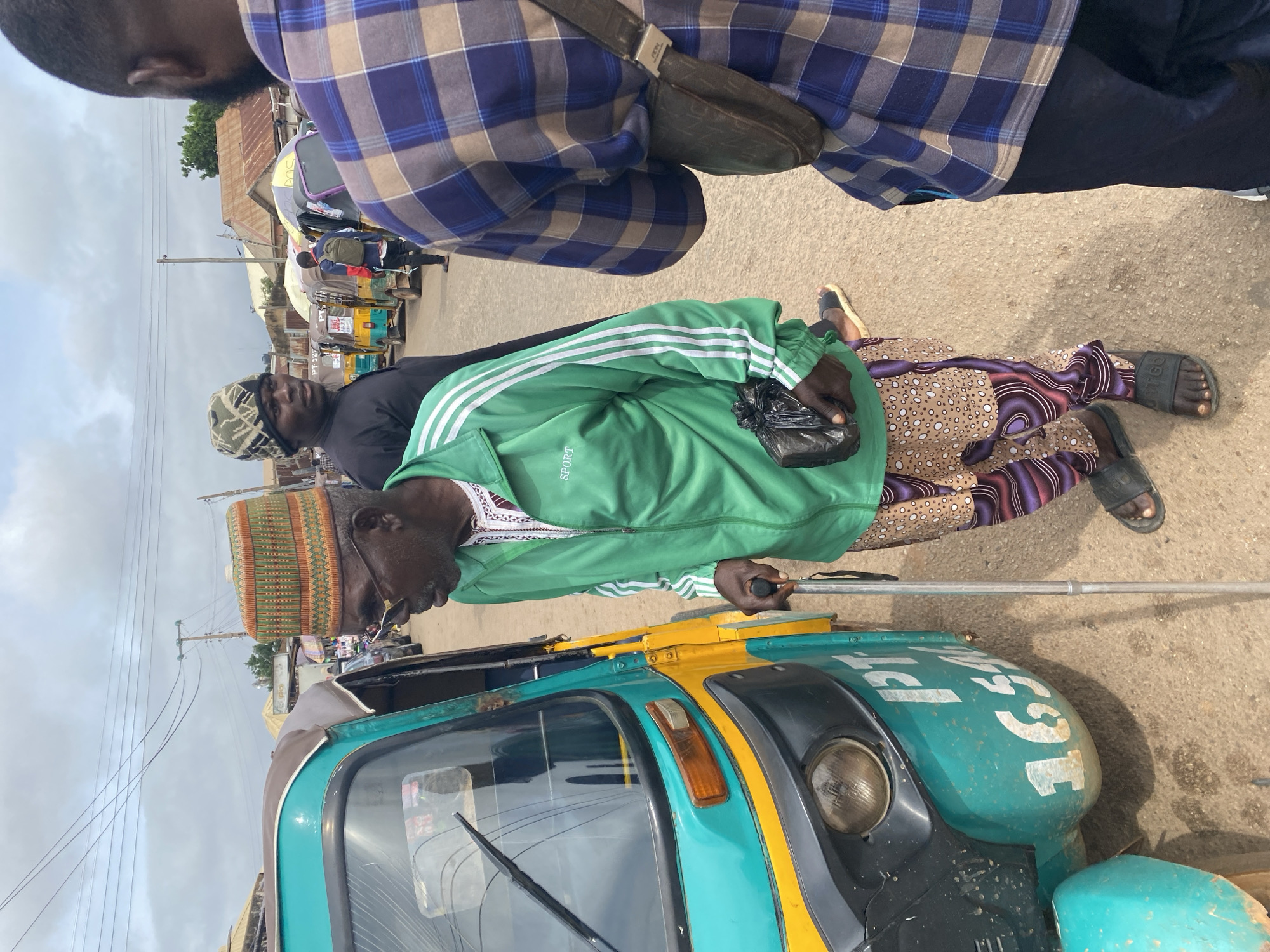In Gura Top, Nigeria, an elderly Black man, wearing a green tracksuit with white stripes, a patterned cap, glasses, and patterned trousers, stands next to a teal and yellow auto-rickshaw. He is holding a black plastic bag in his right hand and a walking stick in his left. The auto-rickshaw has "PT 16546" visible on its side. To the right of the elderly man, another Black man wearing a blue and brown plaid shirt and a dark shoulder bag is partially visible from the back. Behind them, several other auto-rickshaws are parked, and in the background, there are buildings and utility poles under a cloudy sky. The ground is paved with dirt and gravel.