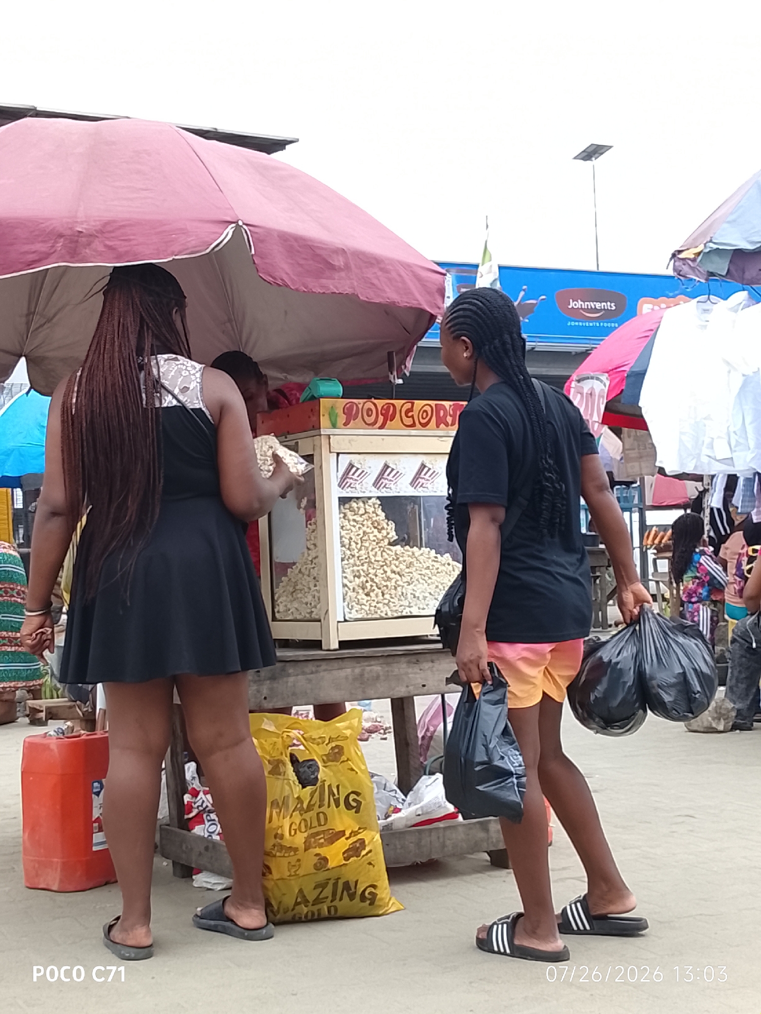 In Lekki, Nigeria, on July 26, 2026, at 13:03, a street scene unfolds beneath a large, pink umbrella. Two young women are the primary subjects. The woman on the left, from behind, wears a black dress with white lace detailing on the shoulders and holds a bag of popcorn. She has long, braided dark hair. The woman on the right, also viewed from behind, is dressed in a black t-shirt and orange shorts. She carries two large black plastic bags, one in each hand. Both women are wearing casual footwear. They are standing in front of a popcorn machine, which is visible with its contents. To the left of the scene, a large orange plastic container is present, and to the right, a yellow bag with text partially visible, possibly indicating "AMAZING GOLD," lies on the ground. Various market stalls and other people are discernible in the background, suggesting a bustling environment. The sky is overcast.