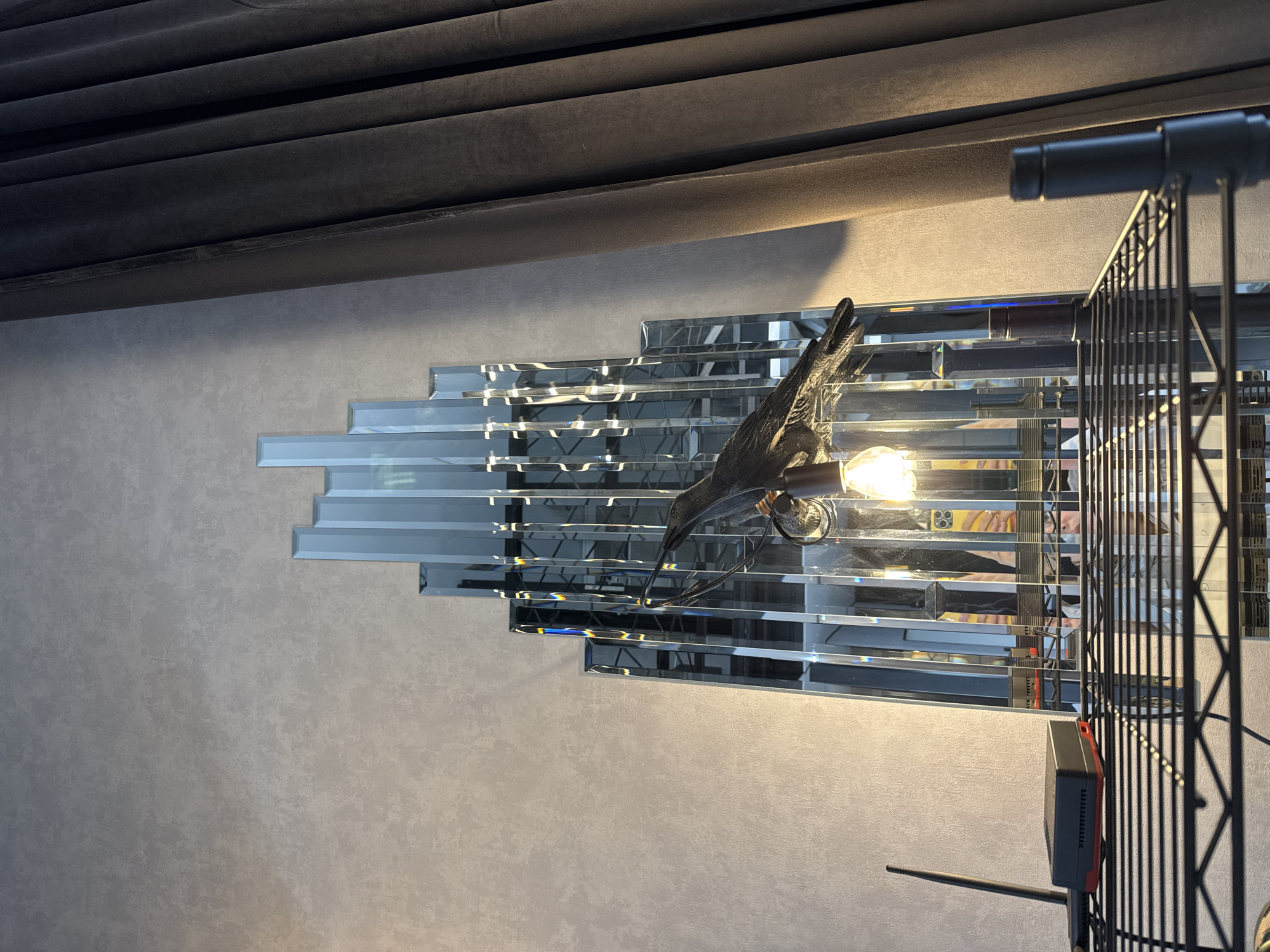 The image captures an interior scene, focusing on a decorative wall installation. Centrally positioned is an art-deco style mirror composed of multiple rectangular, beveled glass segments arranged in a stepped, geometric pattern. Attached to the mirror is a unique light fixture, featuring a dark, bird-shaped element (resembling a hummingbird or small raptor) with its beak pointed towards an exposed, glowing incandescent light bulb. The light bulb illuminates the surrounding area, casting reflections onto the mirror and the textured light grey wall behind it.
In the upper part of the frame, dark, plush curtains or drapes are visible, adding a soft, folded texture to the scene. To the right, a black metal shelving unit or stand with a grid-like design is partially visible. An electronic device, dark in color with a red side panel and an antenna, is mounted on this shelving unit. A small red button on the device is labeled "RESET," and some indistinct white text, possibly part of a brand name, is visible near it. The ambient light suggests an indoor setting, likely in the evening or a dimly lit space, as the artificial light source is prominent. No people are present, and no specific activity is taking place; the image showcases interior decor.