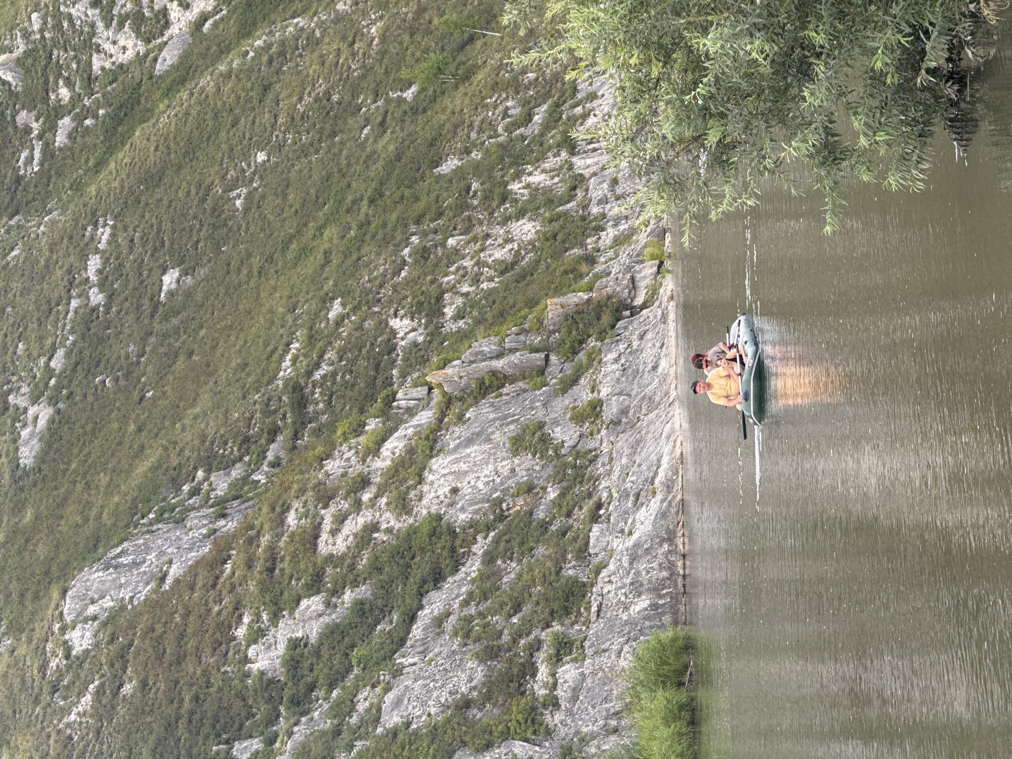 Two people are in a green inflatable boat on a wide, calm river. The person in front is wearing an orange shirt and a dark hat, and is looking towards the right side of the boat. The person behind is wearing a darker shirt and appears to be a child, looking towards the left. The boat is moving from left to right. The riverbanks are lined with tall green grass on the left and a steep, rocky, and vegetated mountainside on the right. The water is a murky brown color and reflects the overcast sky. The background is a large, imposing mountain covered in green vegetation and grey rock formations. This scene appears to be in a natural, rural setting, likely a river valley.
