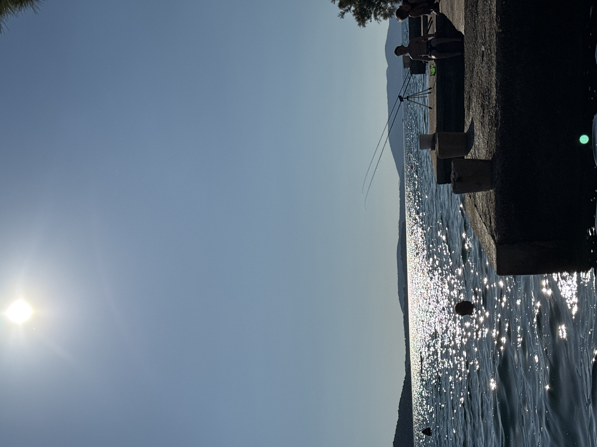 The image depicts a serene scene at the water's edge, likely in Grad Šibenik, Croatia. The sun, positioned on the left side of the frame, casts a bright glare, suggesting it is daytime, possibly late afternoon or early morning due to the soft light. The water is calm, with sunlight sparkling on its surface, creating a shimmering effect. In the background, distant hills or mountains are visible, silhouetted against the pale blue sky.
On a concrete embankment to the right, two individuals are fishing. One person is seated, with fishing rods set up on a tripod. The other person appears to be standing or leaning nearby. Their presence suggests a leisurely activity, enjoying the tranquil environment. A few stones are visible protruding from the water near the embankment, and a small dark object, possibly a buoy or a floating piece of debris, is also present in the water. The overall mood is peaceful and relaxed, characteristic of a sunny day by the sea. No text is visible in the image.