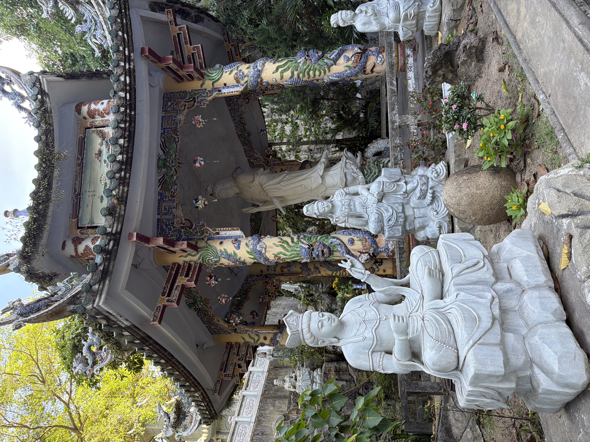 The image shows a courtyard at the Linh Ung Pagoda on Son Tra Peninsula in Da Nang, Vietnam. The central focus is a weathered, grey stone statue of Guanyin, the goddess of mercy, seated in a meditative pose with a crown and intricate jewelry. She is positioned on a tiered stone base. To her left, another grey stone statue of Guanyin is seated on a rocky outcropping, also in meditation. Behind these statues, a large white statue of Guanyin stands prominently within an ornate pavilion. The pavilion itself is decorated with colorful ceramic tiles, dragon motifs, and hanging lanterns. Other smaller statues are visible in the background. The surrounding environment includes lush green trees, foliage, rocks, and paved pathways, indicating an outdoor temple setting. The sun is shining, casting shadows and highlighting the textures of the stone and the vibrant colors of the pavilion.