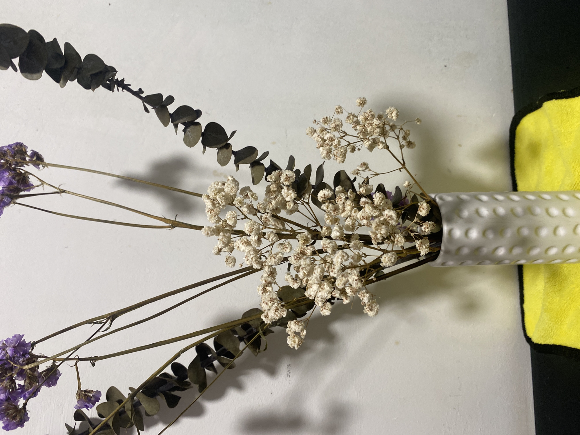 The image shows a close-up, top-down view of a dried floral arrangement in a white vase with a patterned surface. The arrangement includes dried baby's breath (Gypsophila), dried purple statice flowers, and sprigs of dried eucalyptus leaves. The vase is resting on a surface that is partially white and partially covered by a yellow cloth with a black border. The background is a plain white surface.