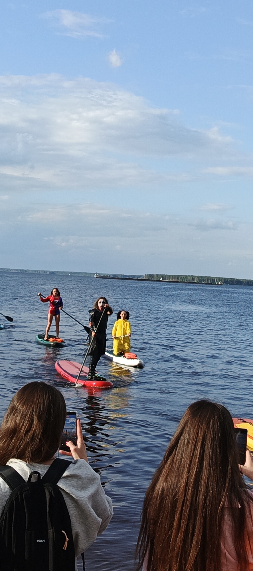 The image shows a group of people on paddleboards in a large body of water, likely a river, in the city of Chaikovsky, Russia. The sky is blue with some scattered clouds. In the foreground, two individuals are visible from the back, looking towards the paddleboarders. One is wearing a grey hoodie and a black backpack, holding a smartphone to take a photo. The other has long brown hair and is also holding a smartphone.
In the middle ground, three people are on paddleboards. The person closest to the foreground is dressed in a black costume with white face paint, holding a paddle. To their left, a person in a red and blue costume is standing on a paddleboard, also holding a paddle. To the right of the person in black, another individual is on a white paddleboard, wearing a bright yellow outfit.
In the background, the far shore of the water is visible, with a line of trees and a large, long barge or ship moving slowly on the water. The overall scene depicts an outdoor recreational activity.