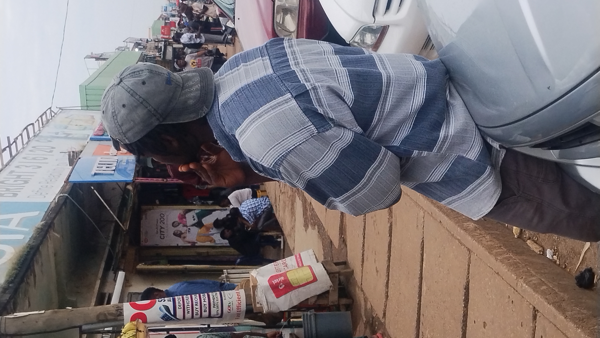 In an outdoor urban setting, a Black male wearing a grey baseball cap and a blue and white striped long-sleeved shirt sits on the hood of a silver car. He is facing away from the camera, with his left hand touching his face and his right hand holding a cigarette to his mouth. He is leaning against a brick building with various signs. One sign above the building reads "BRIGHT 3/16/120". To the left of this sign is another sign that reads "TELCOM". A red and white "Airtel" branded bag is leaning against a wooden post in front of the building. In the background, there are other people and vehicles, indicating a busy street. The ground is paved with concrete.