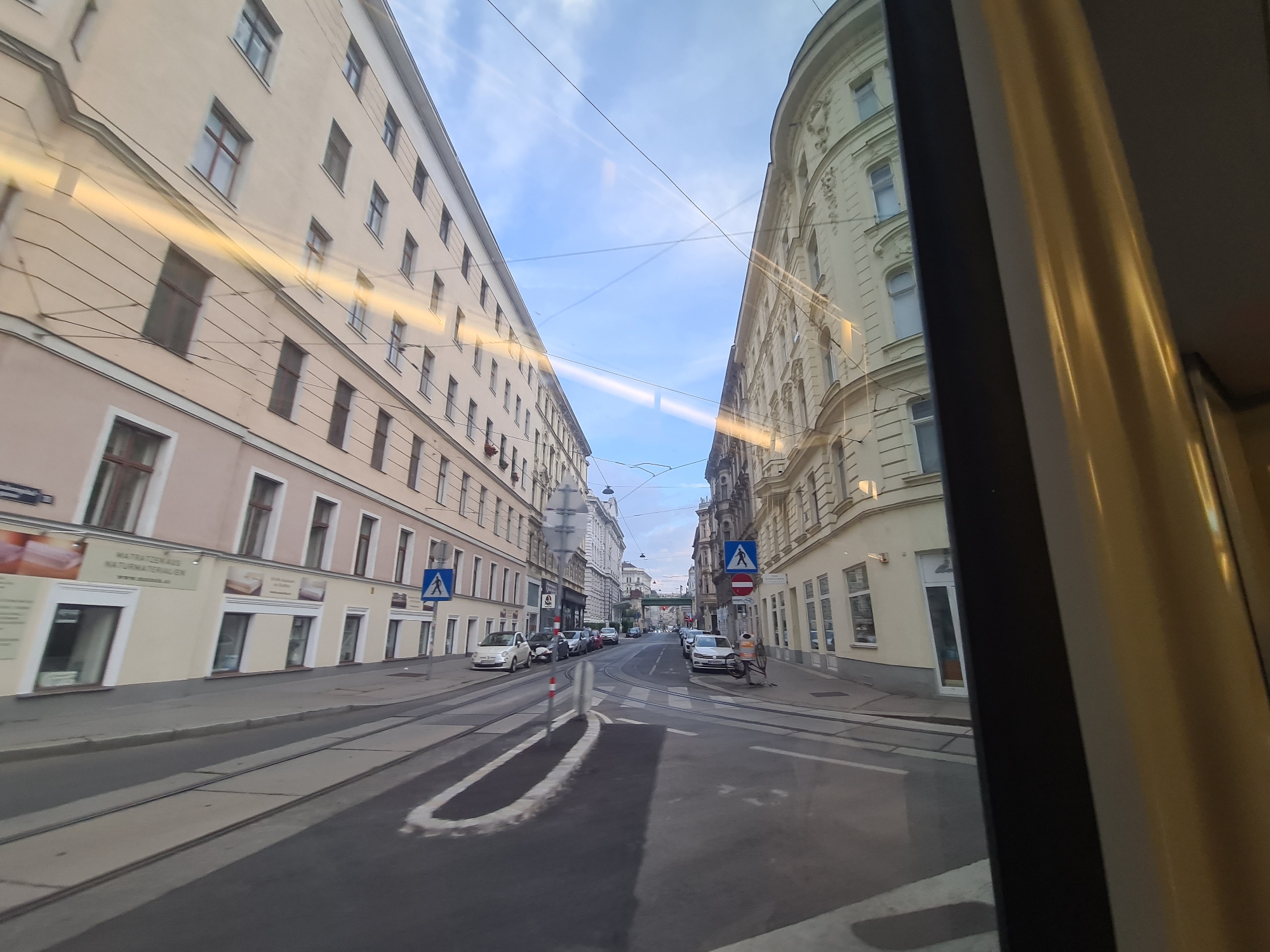 The image shows a view from a tram window looking down a street in Vienna, Austria. The street is lined with buildings on both sides, characteristic of European city architecture, with ornate facades on the right and simpler, taller apartment buildings on the left. Tram tracks are visible in the road, indicating active public transport. Several cars are parked along the street. Pedestrian crossing signs are visible, one with a blue background and a white walking figure, and another with a red circle and a diagonal line over a walking figure, implying a prohibition. The sky is cloudy but bright, suggesting daytime. There are overhead wires, likely for the tram system. The reflections on the window suggest the photograph was taken from inside a moving vehicle.